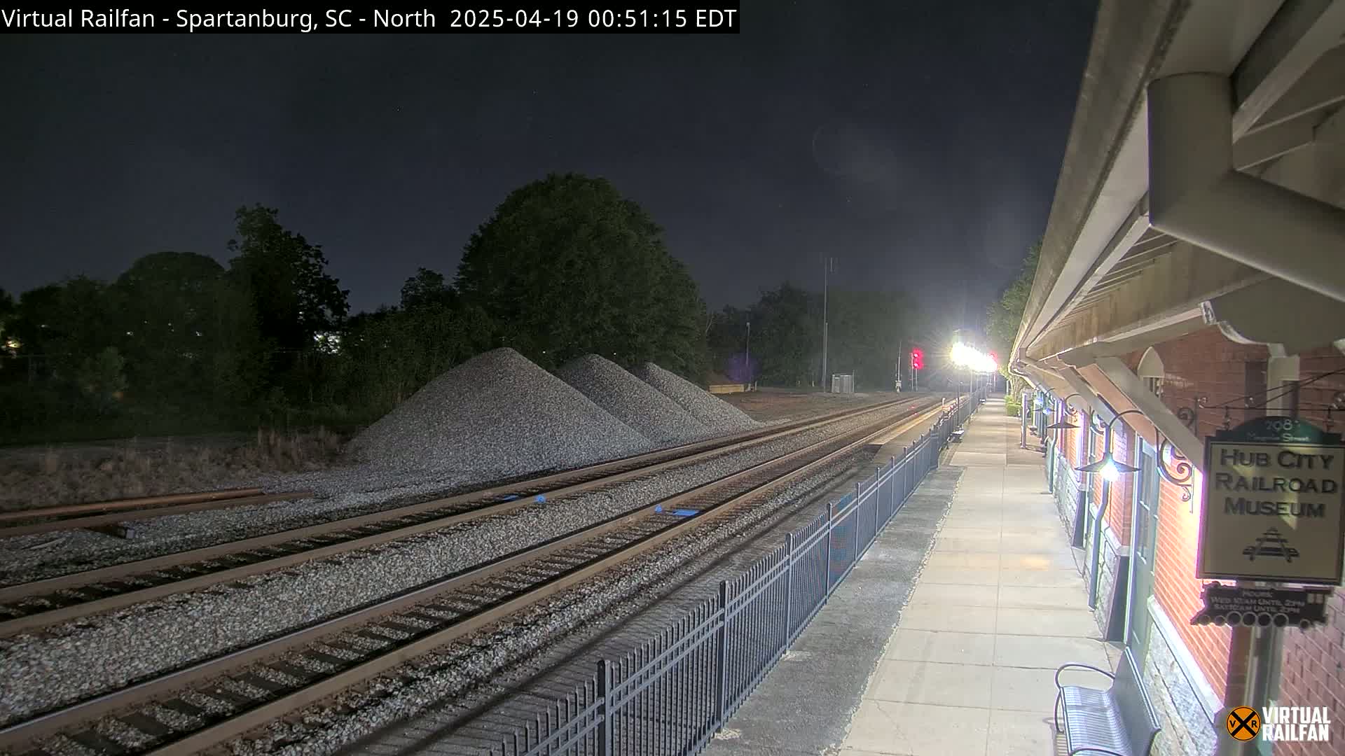 Under a dark night sky, train tracks extend from a nighttime train station towards piles of gravel, bordered by a metal fence.