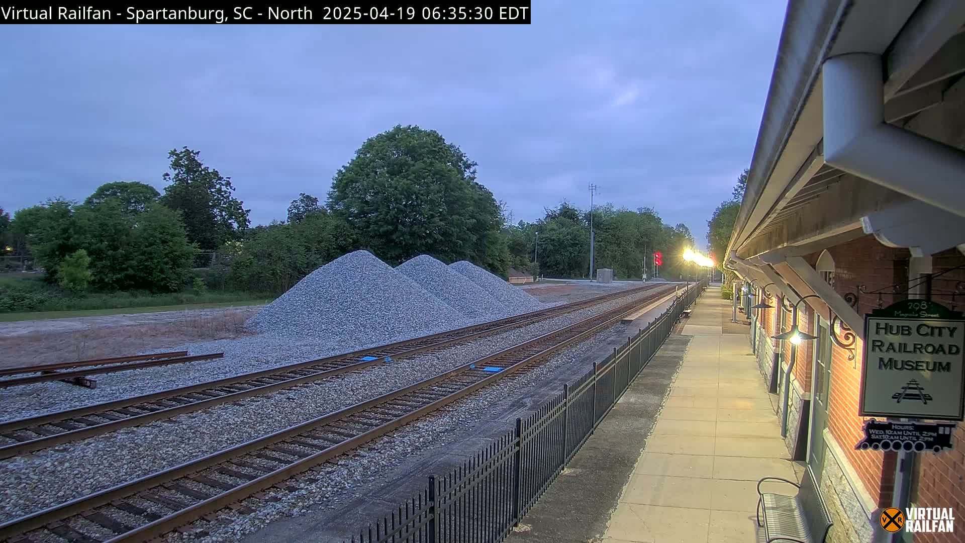 Under a twilight sky, railroad tracks extend from a train platform past piles of gravel toward a line of trees.