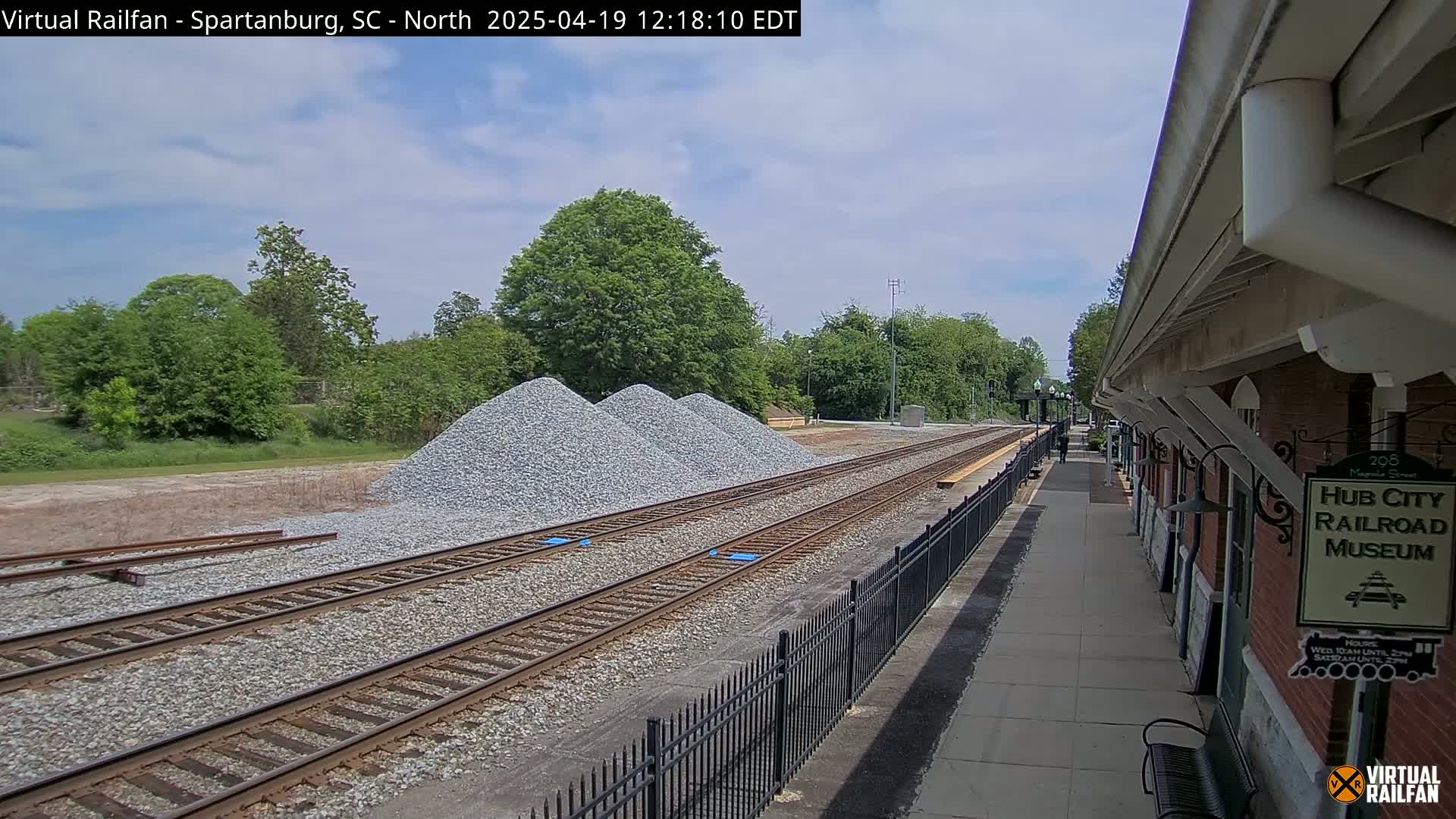 Under a partly cloudy sky, railroad tracks extend from a train station past several piles of gravel toward a line of trees.