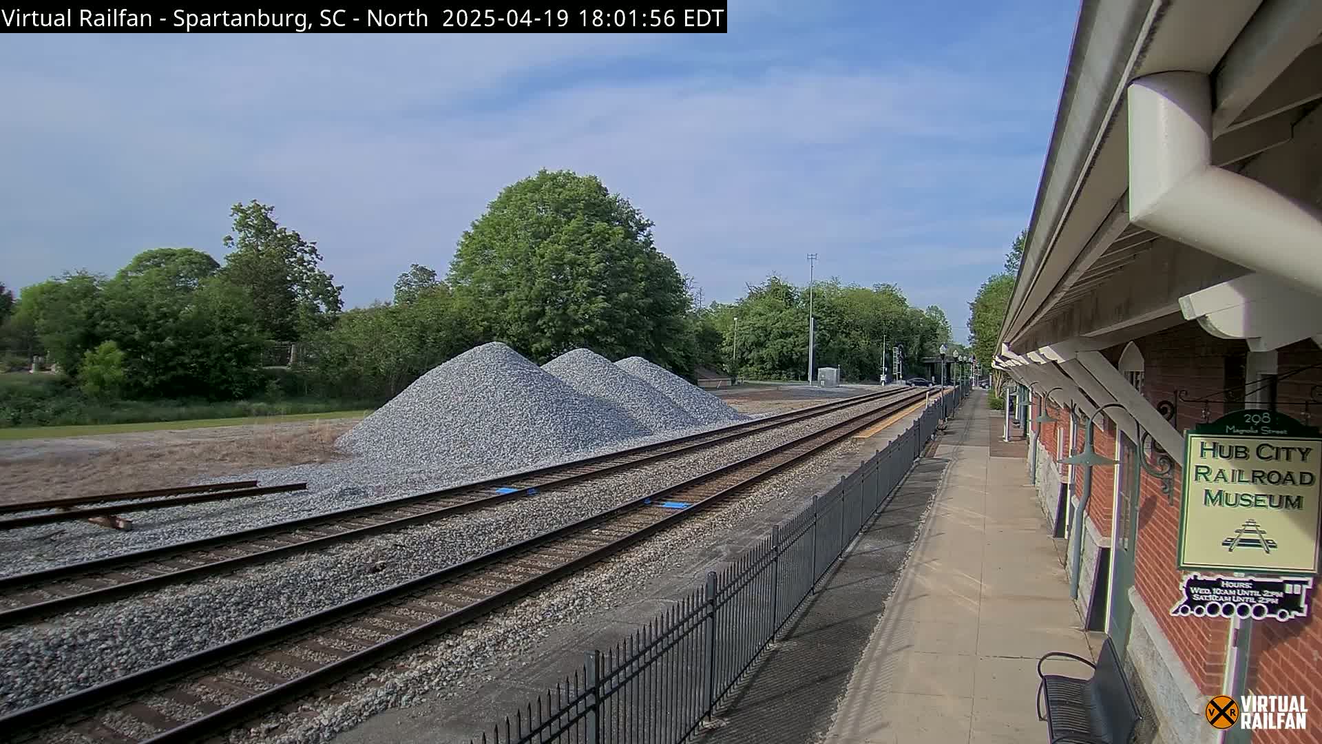 Several piles of gravel are next to railroad tracks that lead to a brick railroad museum under a partly cloudy sky.