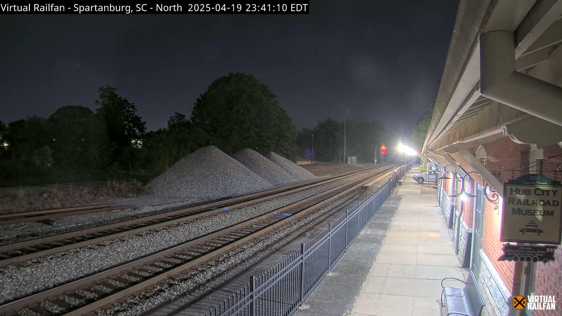 Under a dark night sky, train tracks extend from a nighttime train station, past piles of gravel toward a wooded area.