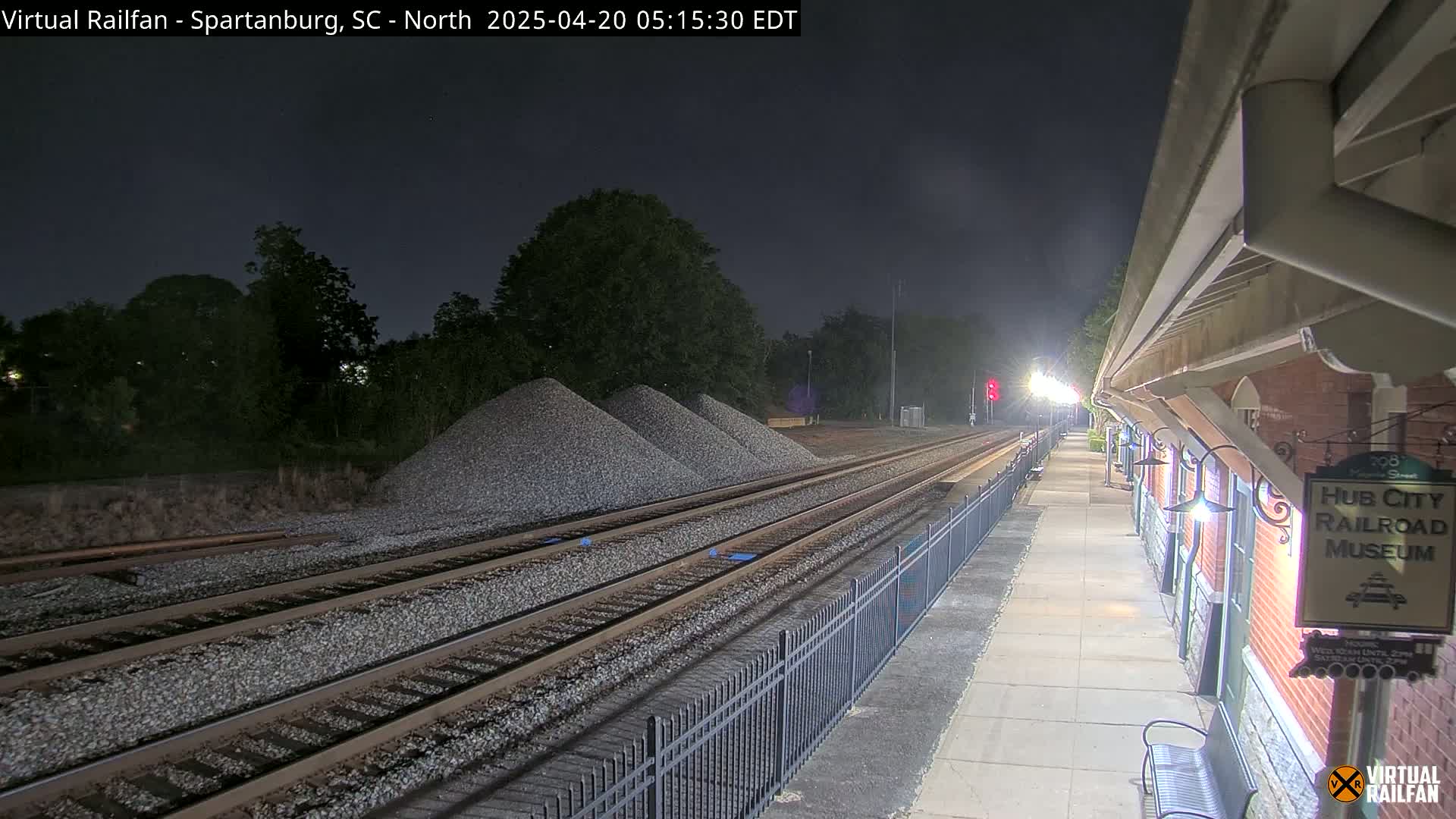Under a dark night sky, railroad tracks extend from a train platform alongside a brick building, with piles of gravel visible nearby.
