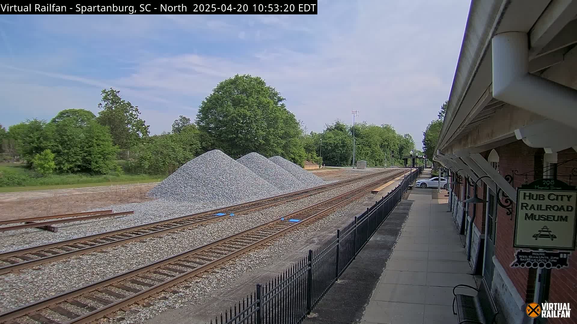 Under a partly cloudy sky, railroad tracks extend from a train station toward three piles of gravel next to some greenery.
