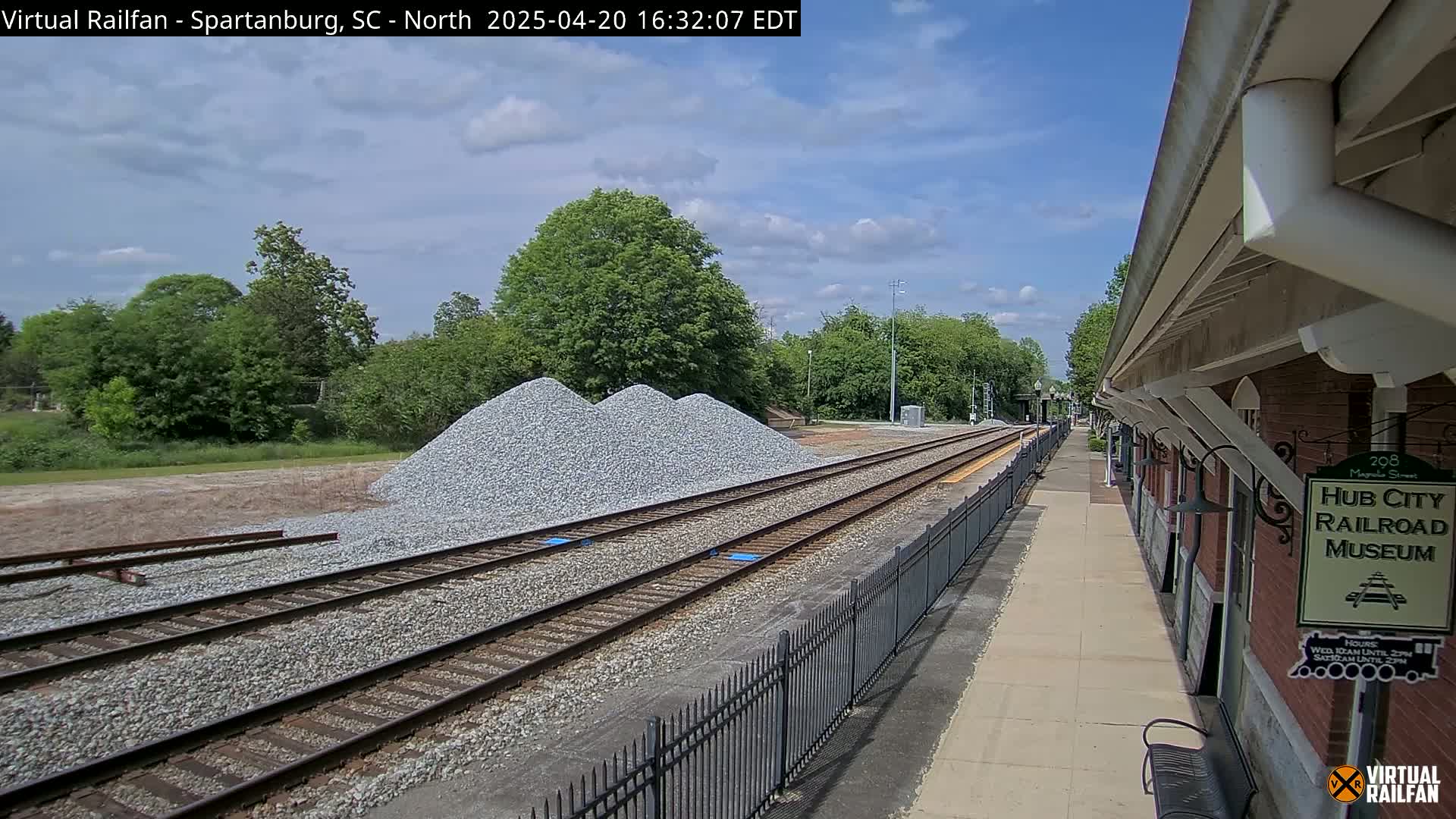 A partly cloudy sky overlooks railroad tracks next to a brick building, with piles of gravel nearby.