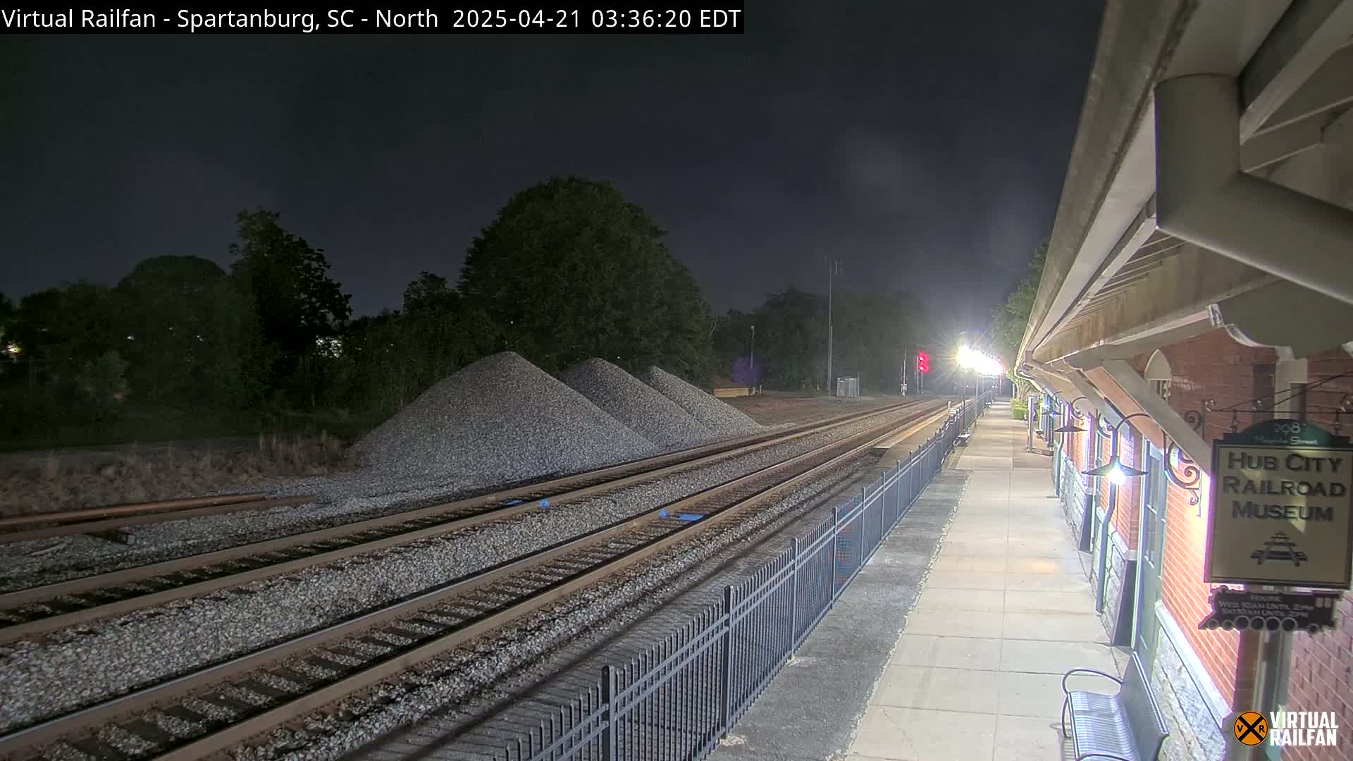 At night, train tracks extend from a station platform towards piles of gravel next to a line of trees under a clear, dark sky.
