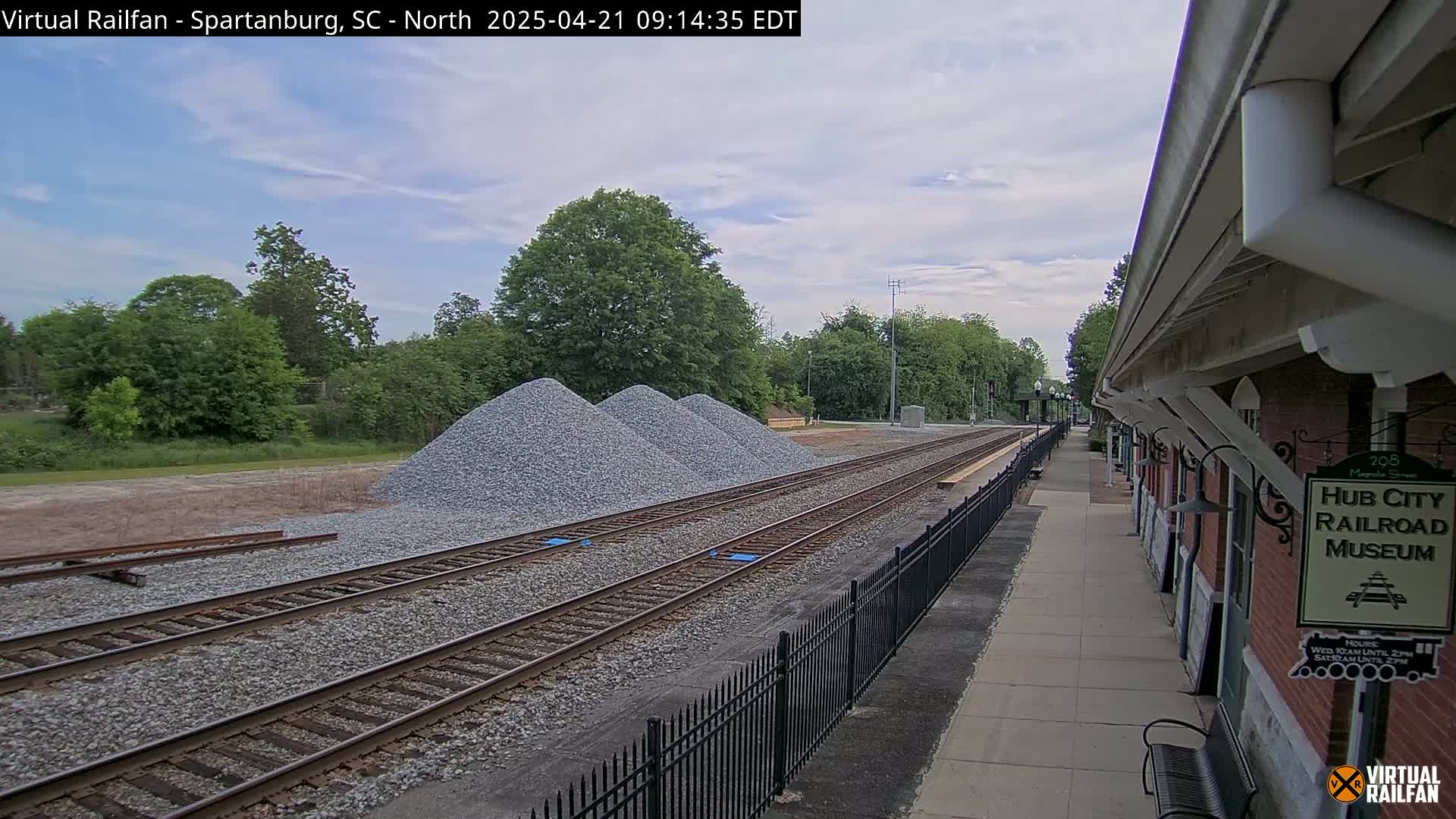 A partly cloudy day reveals railroad tracks next to a brick building, with several piles of gravel nearby.