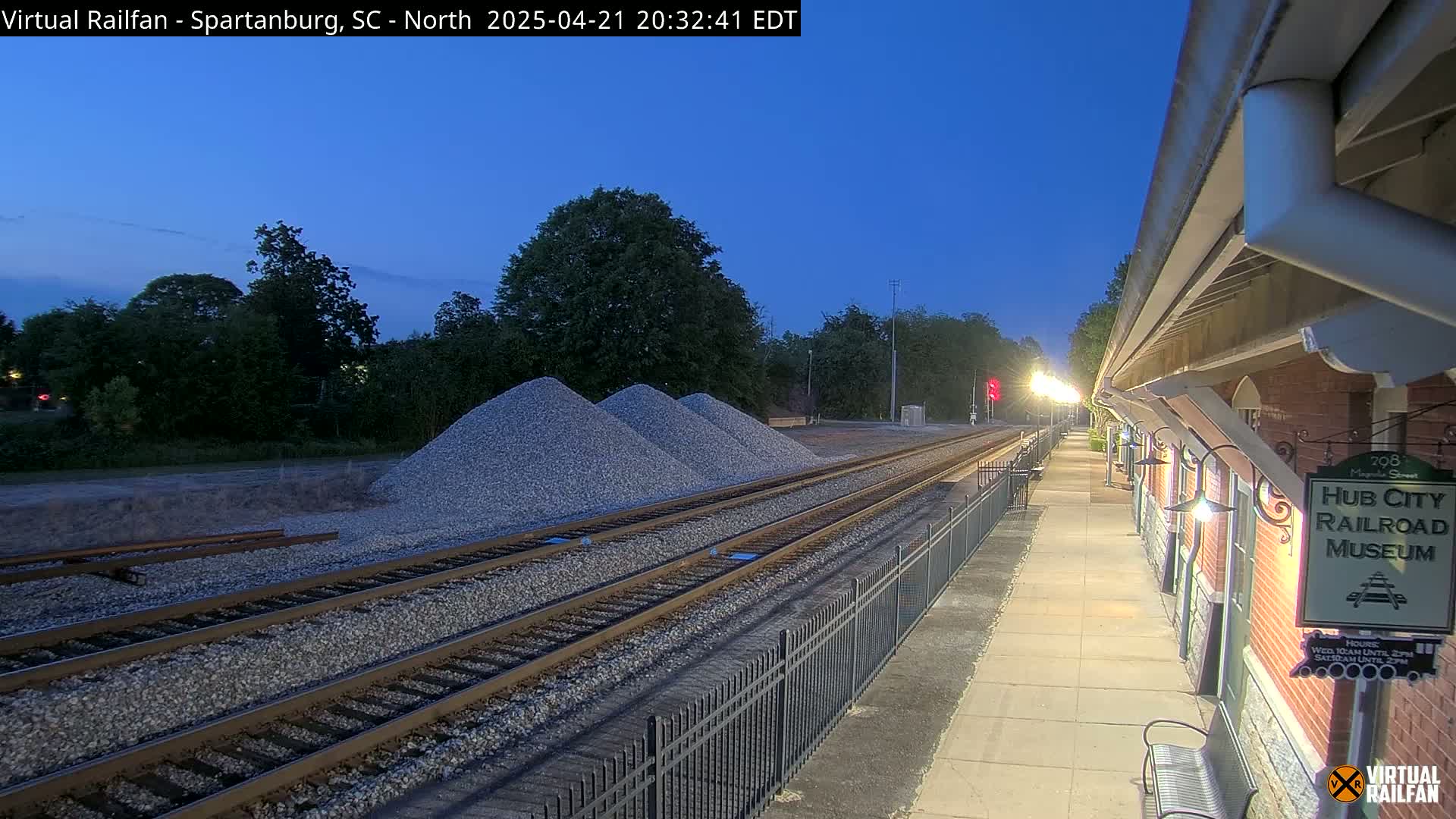 Under a twilight sky, railroad tracks extend from a train platform toward piles of gravel next to a brick building.