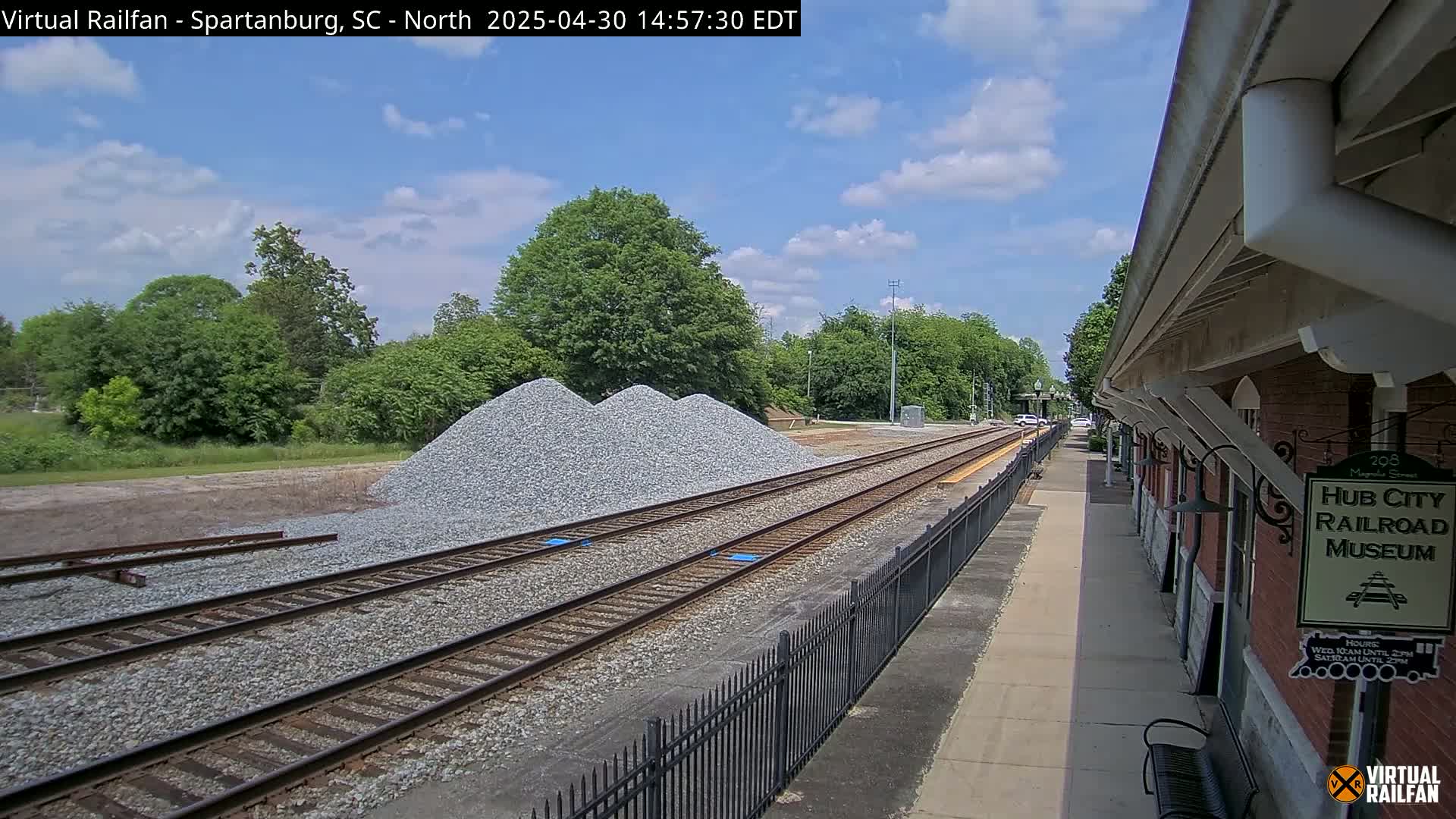 A partly cloudy day shows railroad tracks leading to a brick railroad museum, alongside piles of gravel and lush green trees.