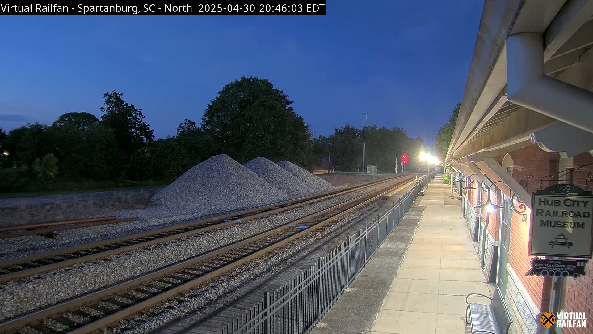 Under a twilight sky, railroad tracks extend from a train platform beside a brick building toward piles of gravel and a line of trees.
