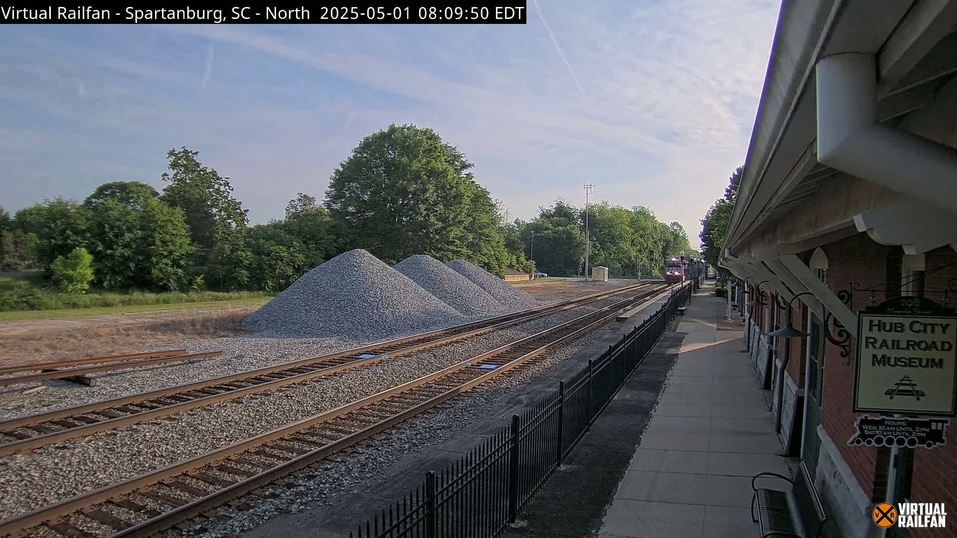 Under a partly cloudy sky, a train passes by a railroad platform next to several piles of gravel.