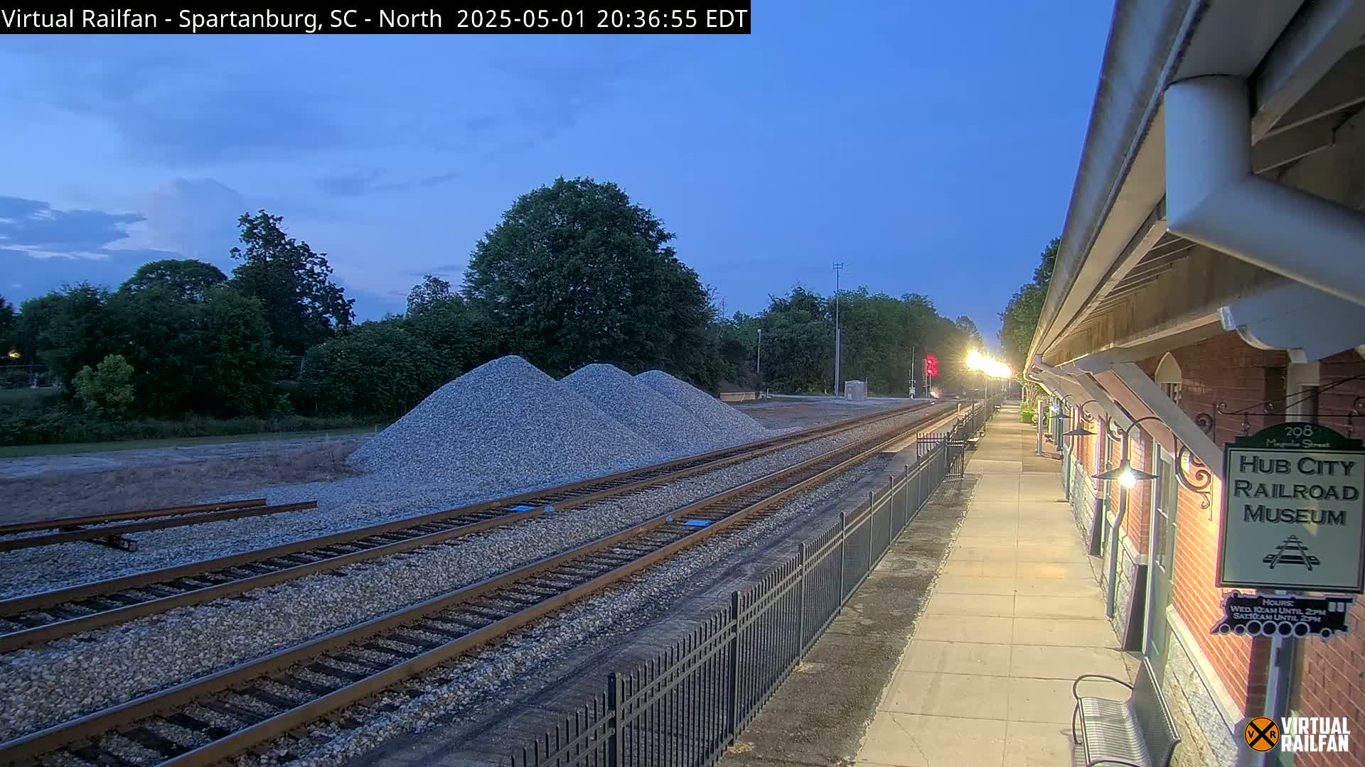 Under a twilight sky, railroad tracks extend from a train platform past piles of gravel toward a line of trees.