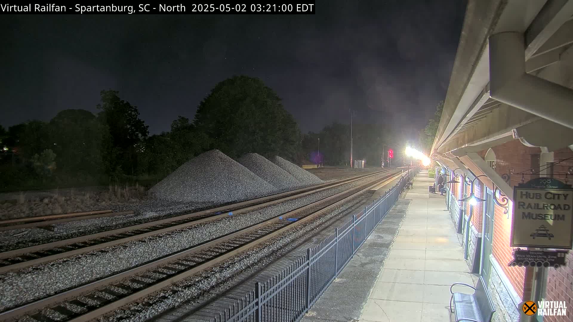 At night, a train station platform next to railroad tracks is illuminated, with piles of gravel nearby.