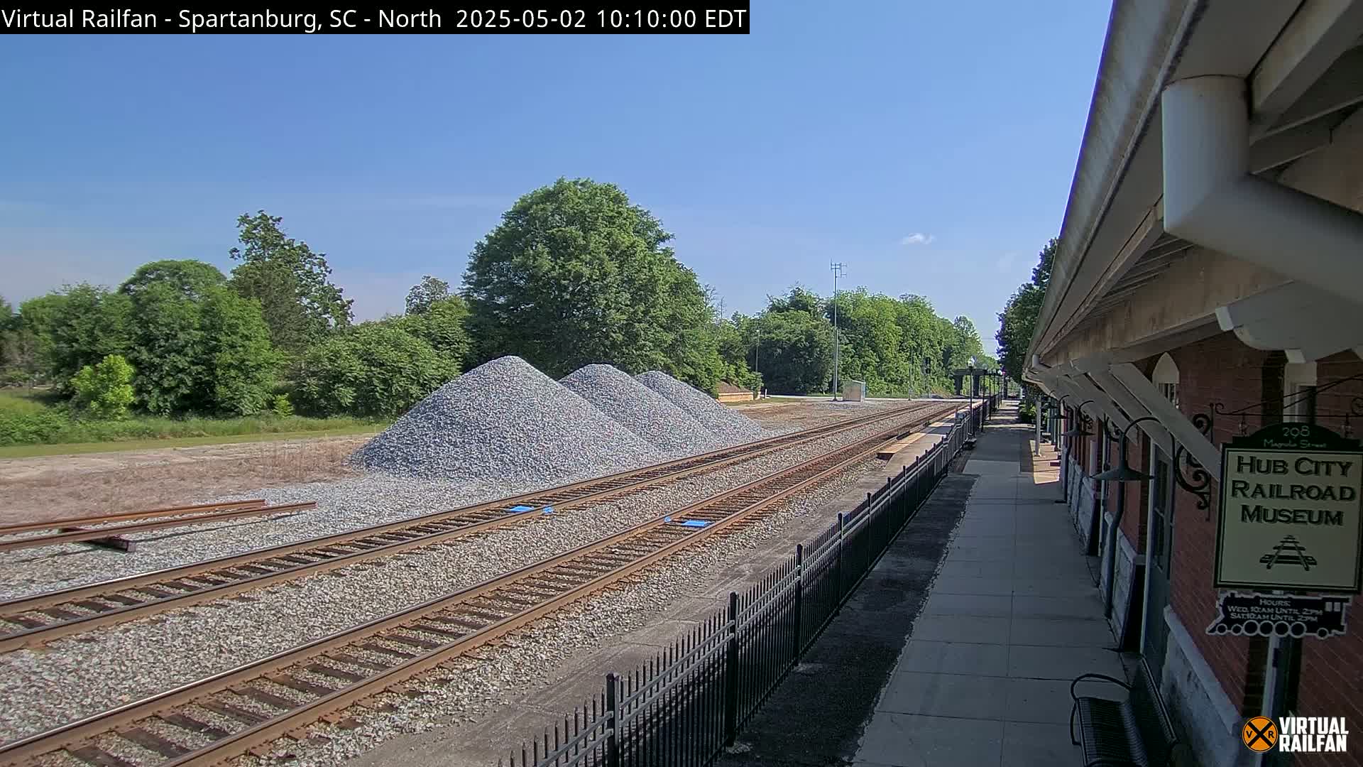 Three piles of gravel sit beside railroad tracks next to a brick building under a clear blue sky.