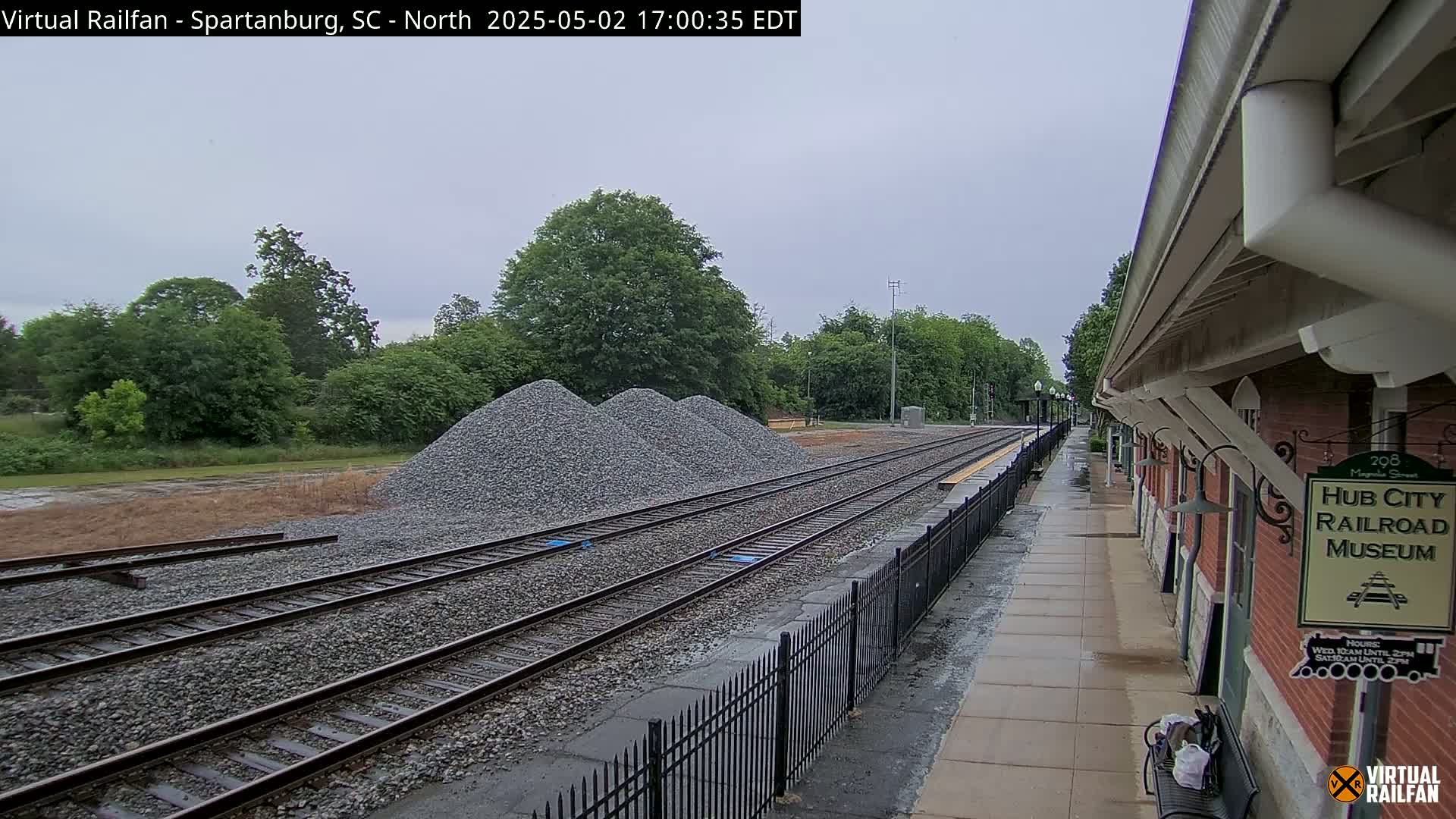 Several piles of gravel sit beside railroad tracks next to a brick building under an overcast sky.