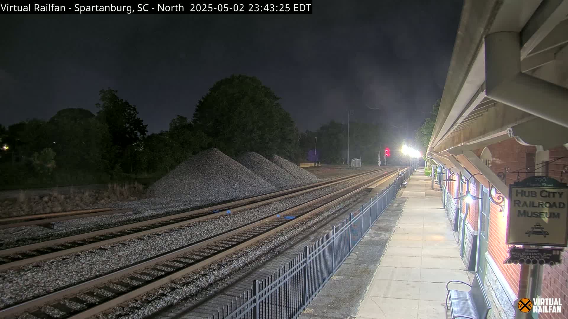 A nighttime view of train tracks extending into the distance, beside a brick building and piles of gravel under a dark sky.