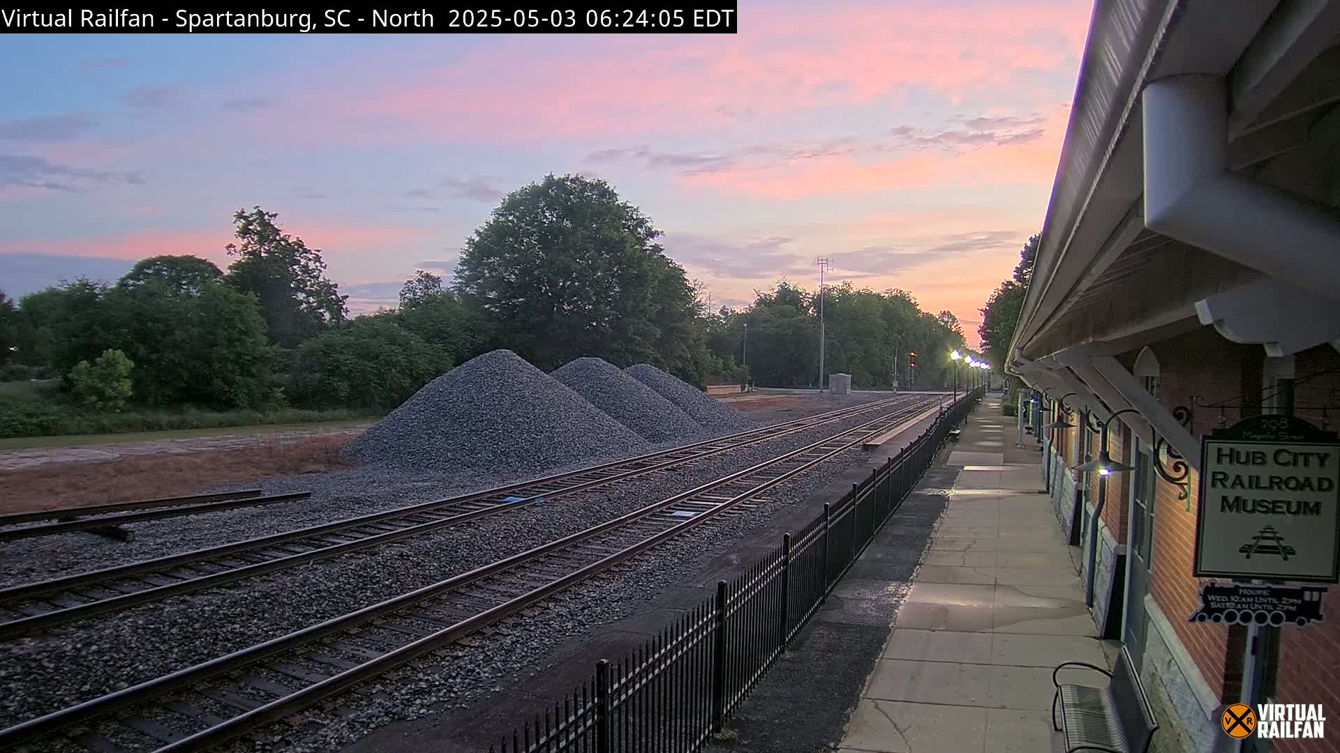 Under a pinkish sunrise, railroad tracks extend from a brick building alongside a paved walkway, with several piles of gravel nearby.
