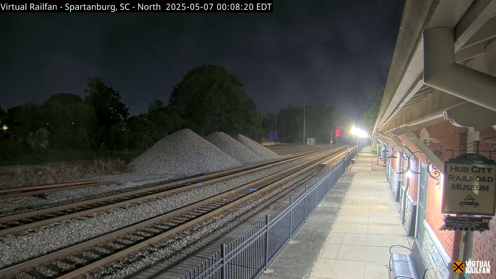 At night, a train platform illuminated by lights is next to railroad tracks with piles of gravel nearby.