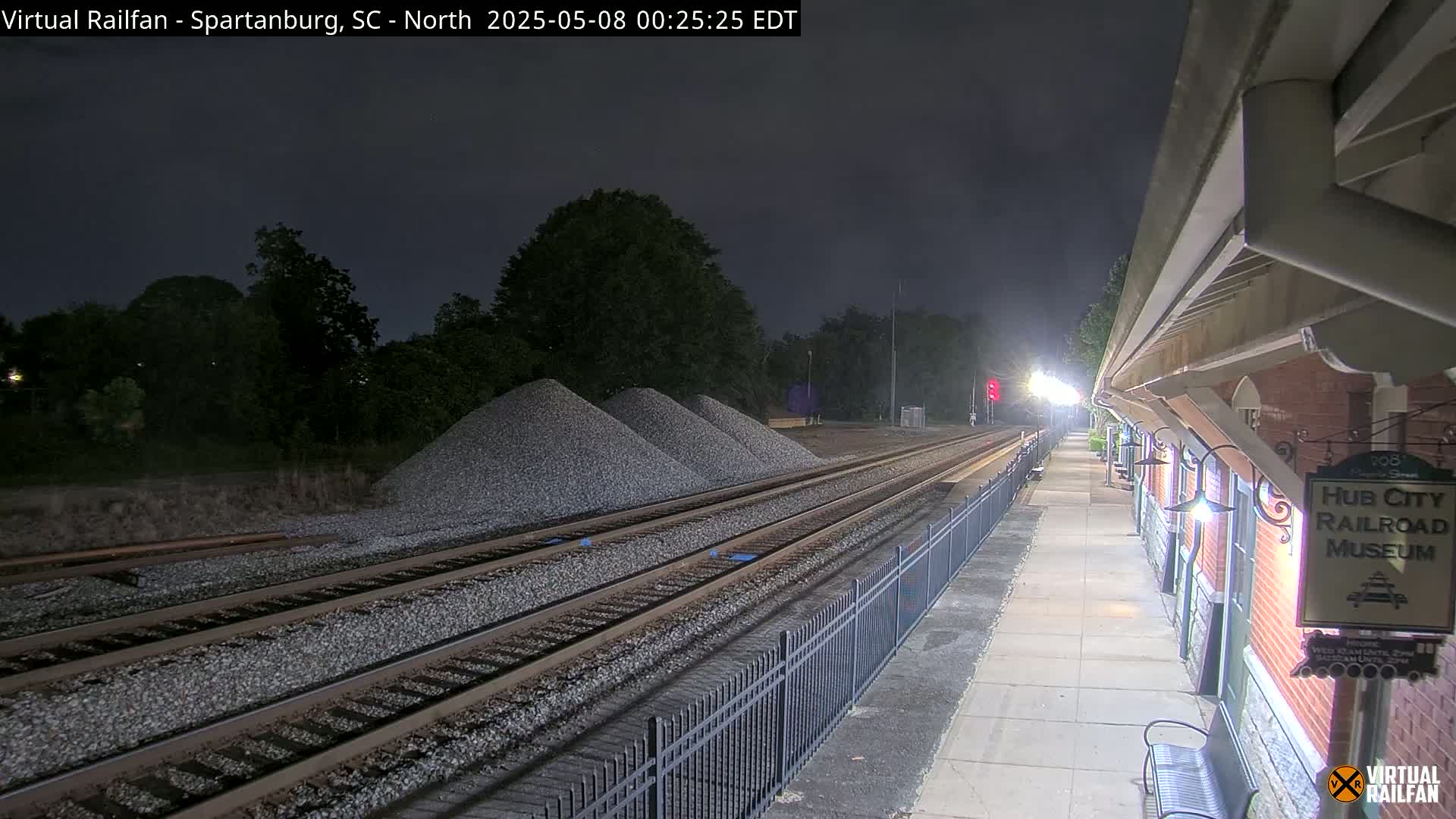 Under a dark night sky, railroad tracks extend toward a pile of gravel near a brick building with lights illuminating a walkway.