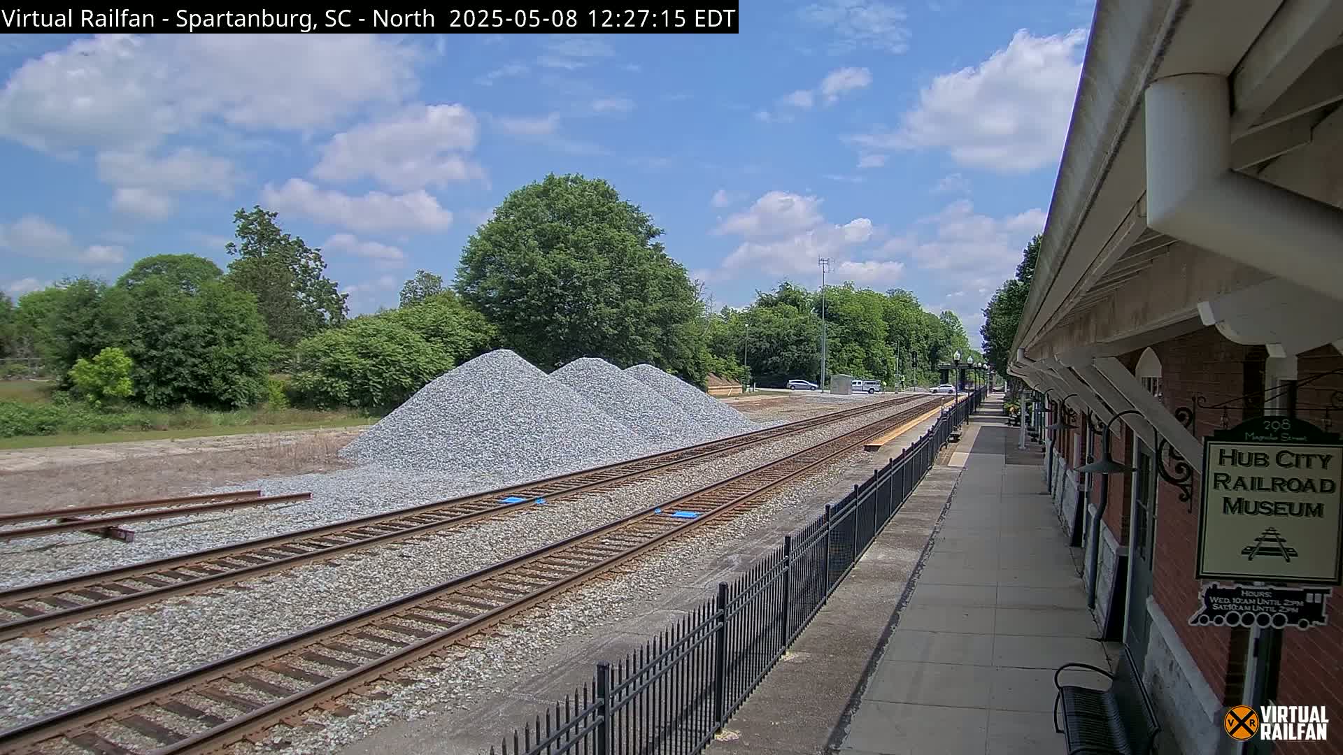 Under a partly cloudy sky, railroad tracks with piles of gravel alongside them extend toward a brick building next to a paved walkway.