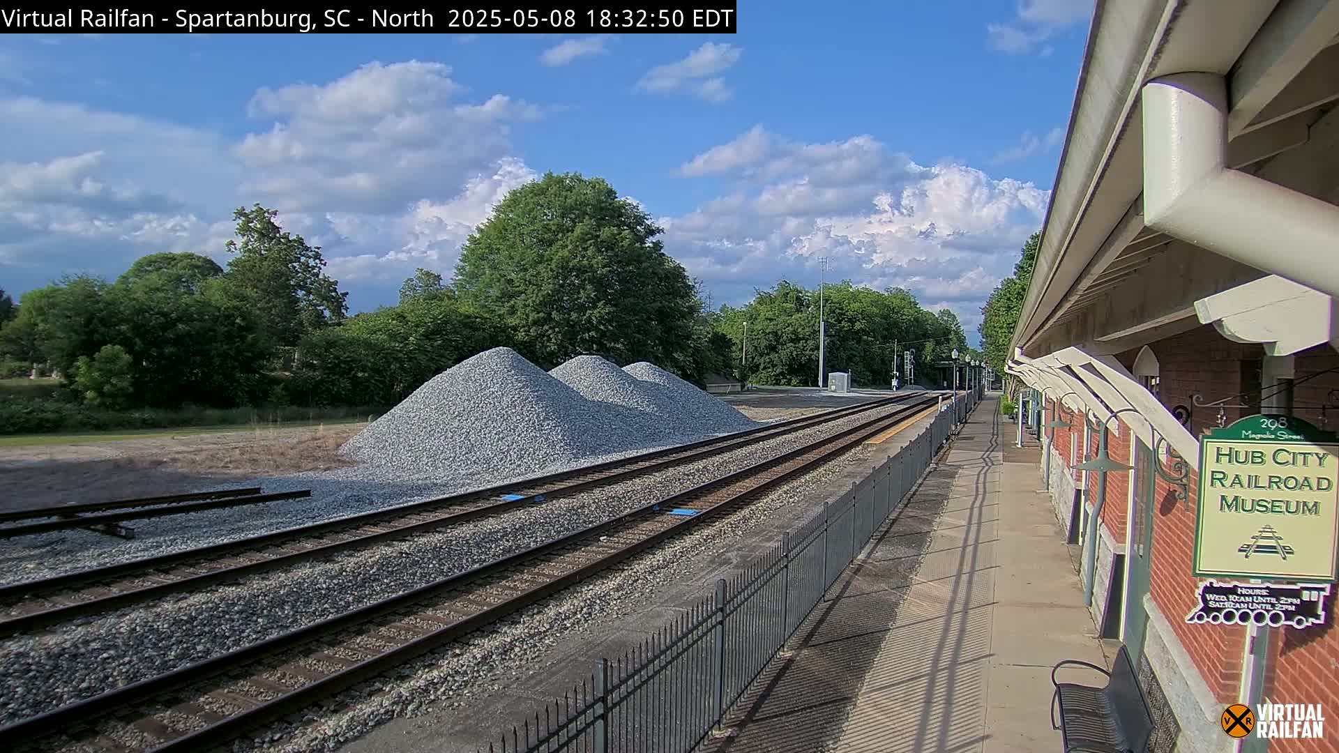 Under a partly cloudy sky, railroad tracks extend from a train platform beside piles of gravel toward a line of trees.
