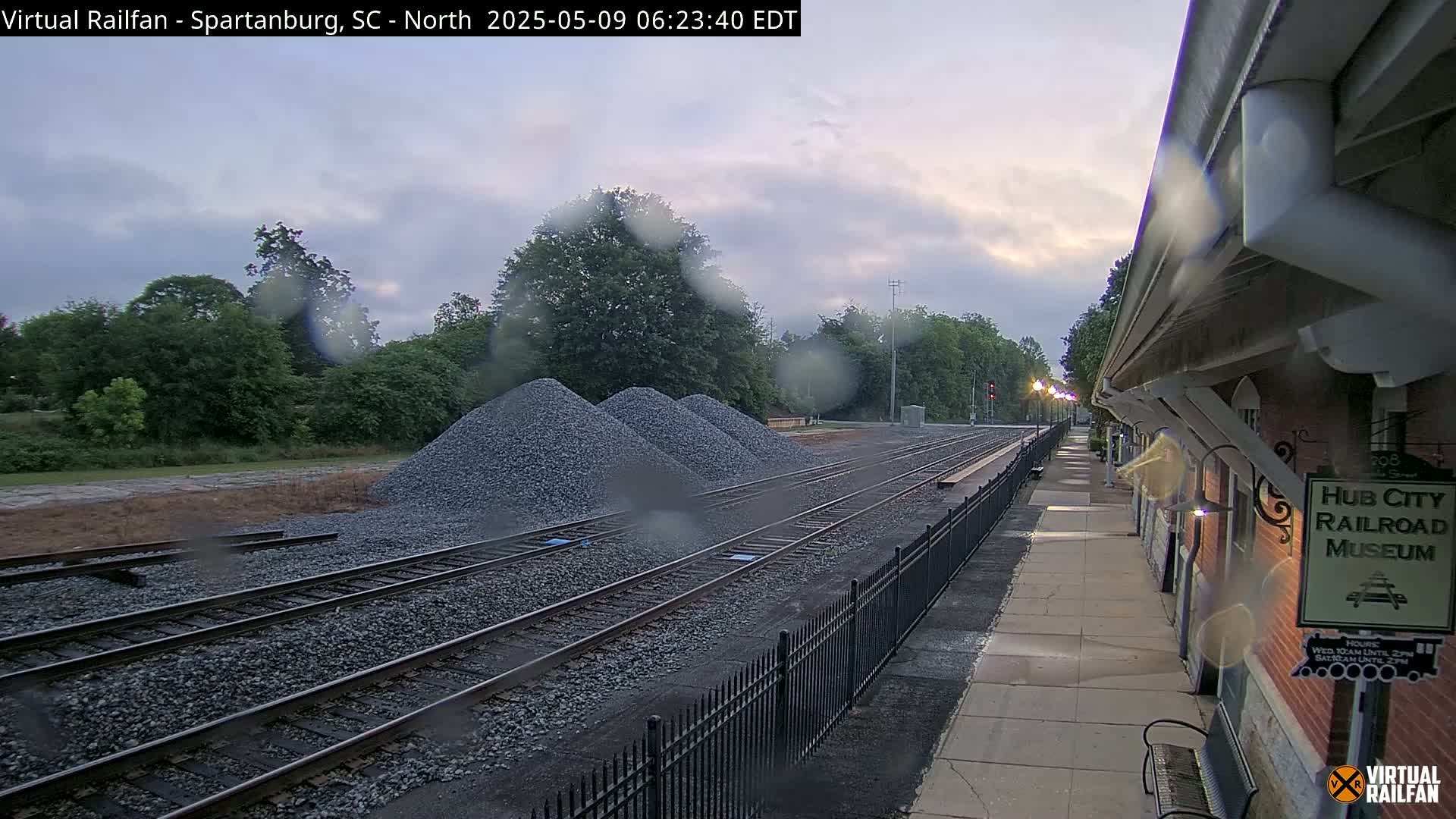 Rain-covered railroad tracks and piles of gravel are visible near a train station platform on an overcast morning.
