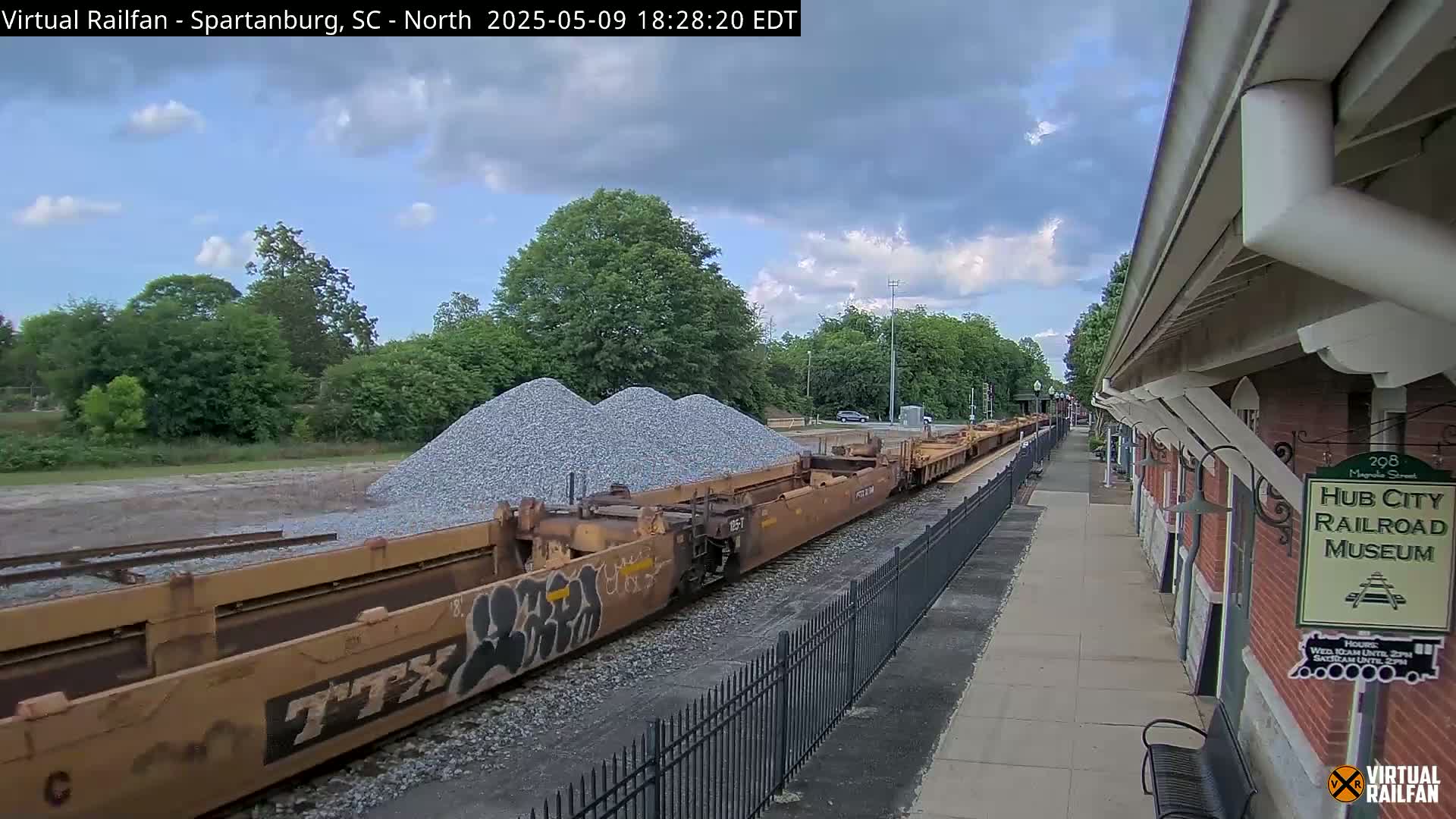 A partly cloudy day shows a freight train carrying piles of gravel alongside a railroad museum.