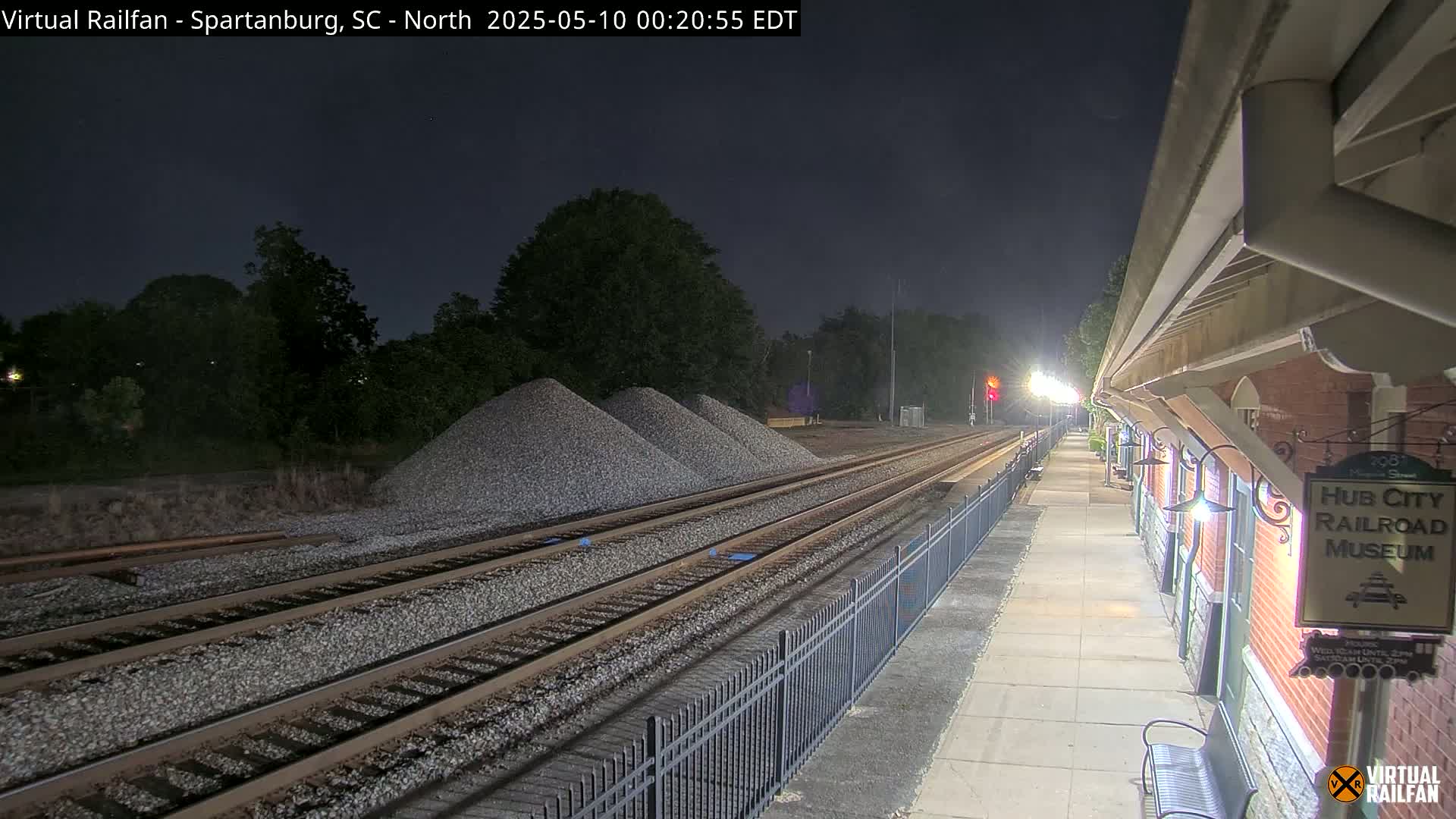 At night, railroad tracks with piles of gravel beside them extend towards a brick building next to a paved walkway, under a dark sky.