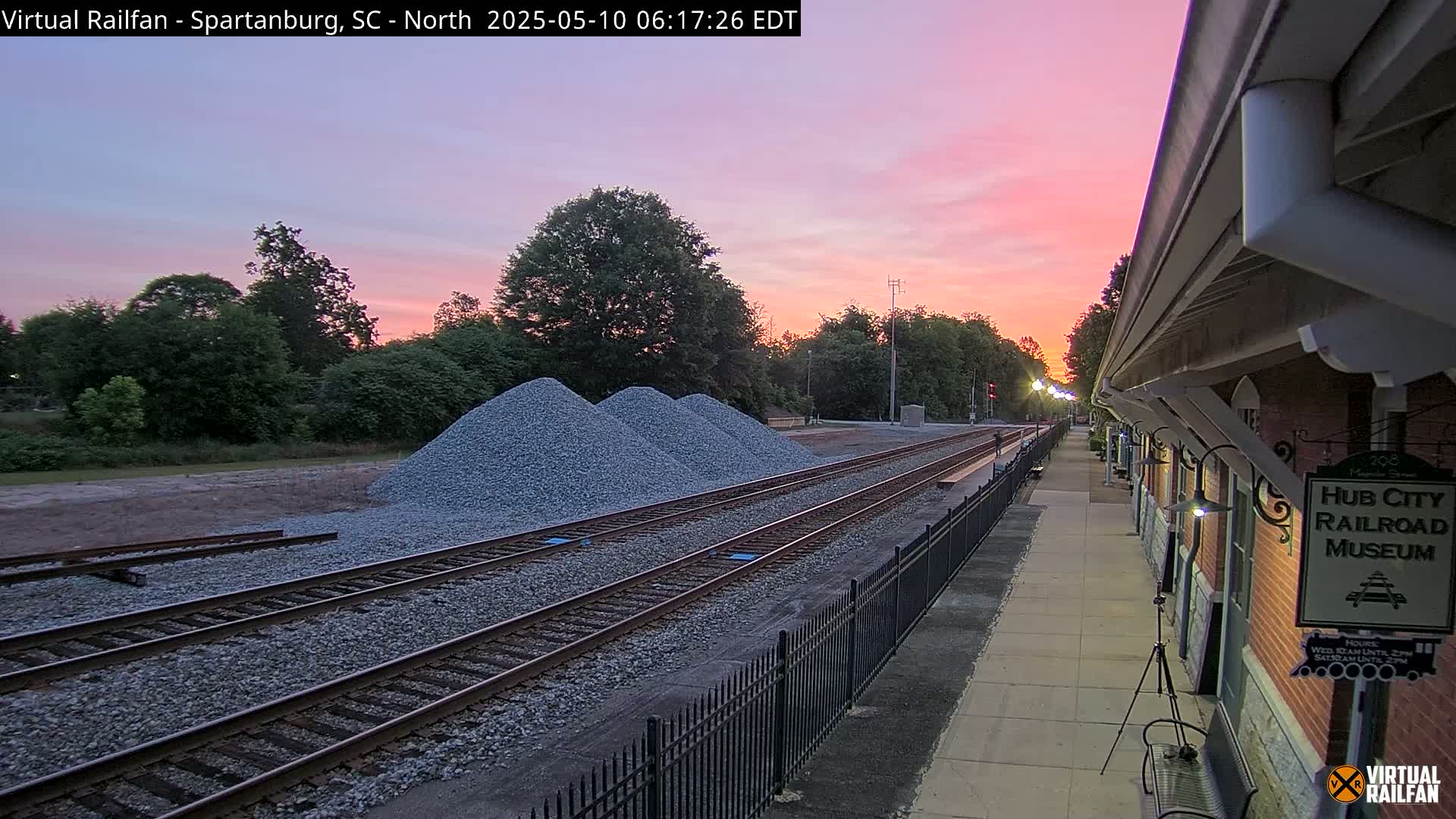 A train track with three piles of gravel next to it runs alongside a brick railroad museum building at sunset.