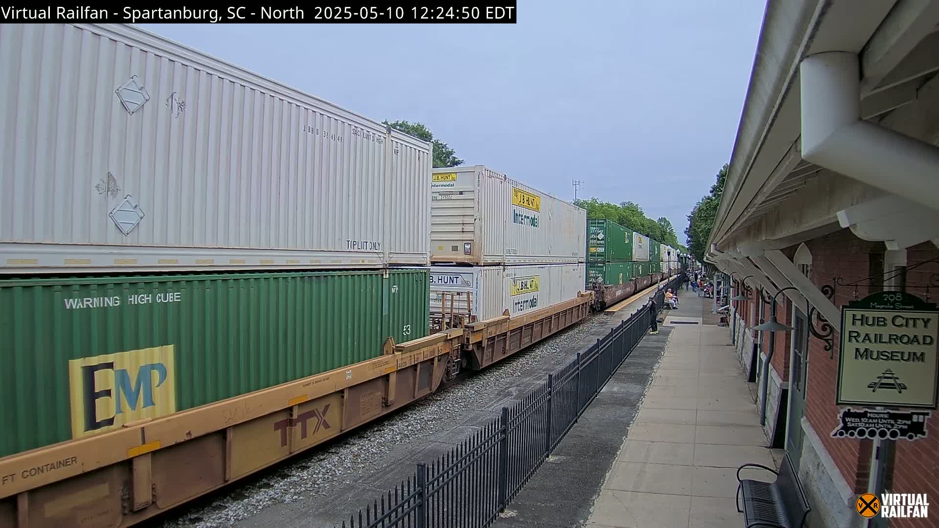 A freight train carrying stacked shipping containers passes by a brick railroad museum on an overcast day.
