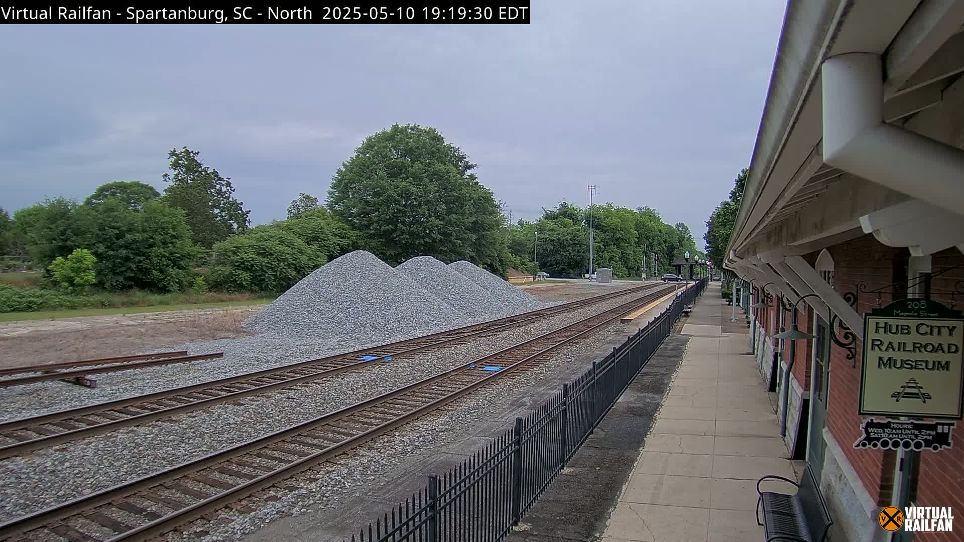 A railroad track runs alongside a brick building with a small sign and several piles of gravel under an overcast sky.