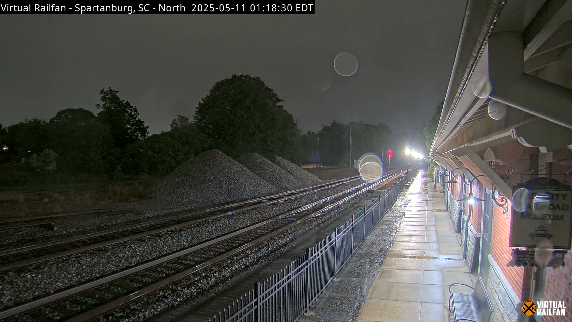 A nighttime view of train tracks next to a brick building under rainy conditions, with piles of gravel nearby.