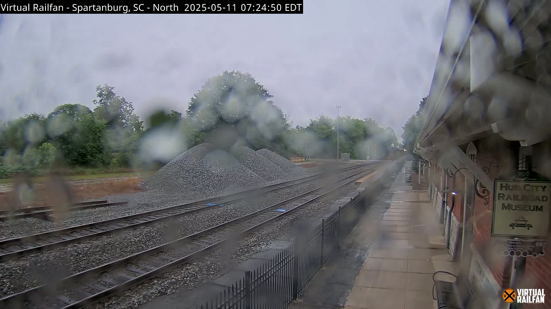 Rain falls on a railway platform with train tracks and piles of gravel visible through a rain-streaked window.