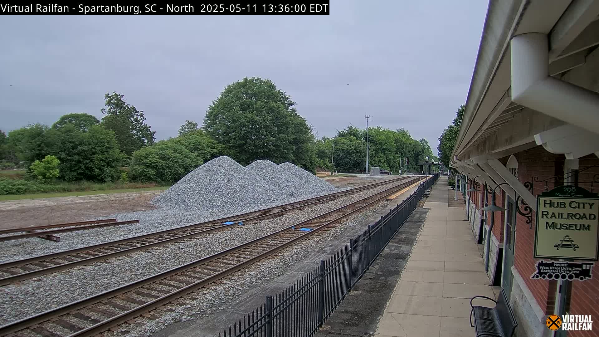 Several piles of gravel sit beside train tracks next to a brick railroad museum building on an overcast day.