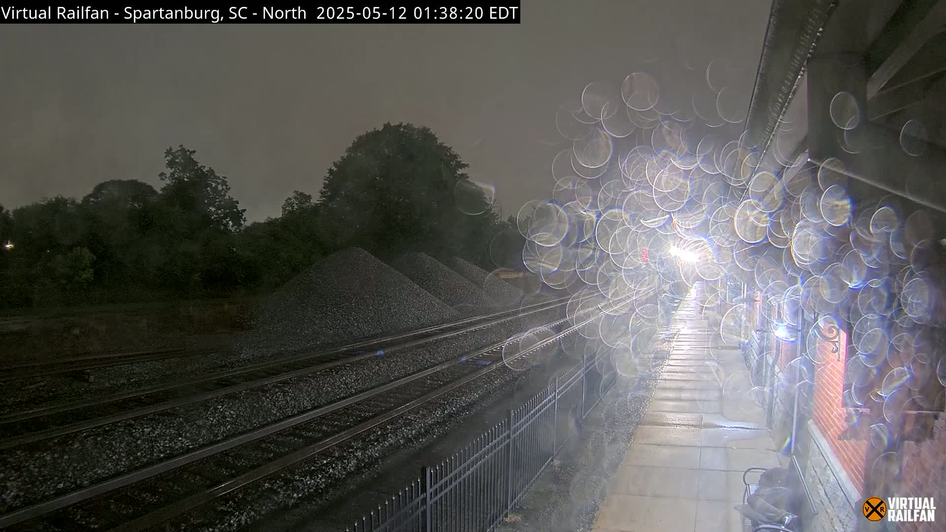 A nighttime view of rain-covered train tracks next to a platform, with piles of gravel in the background and blurry lights from an approaching train.