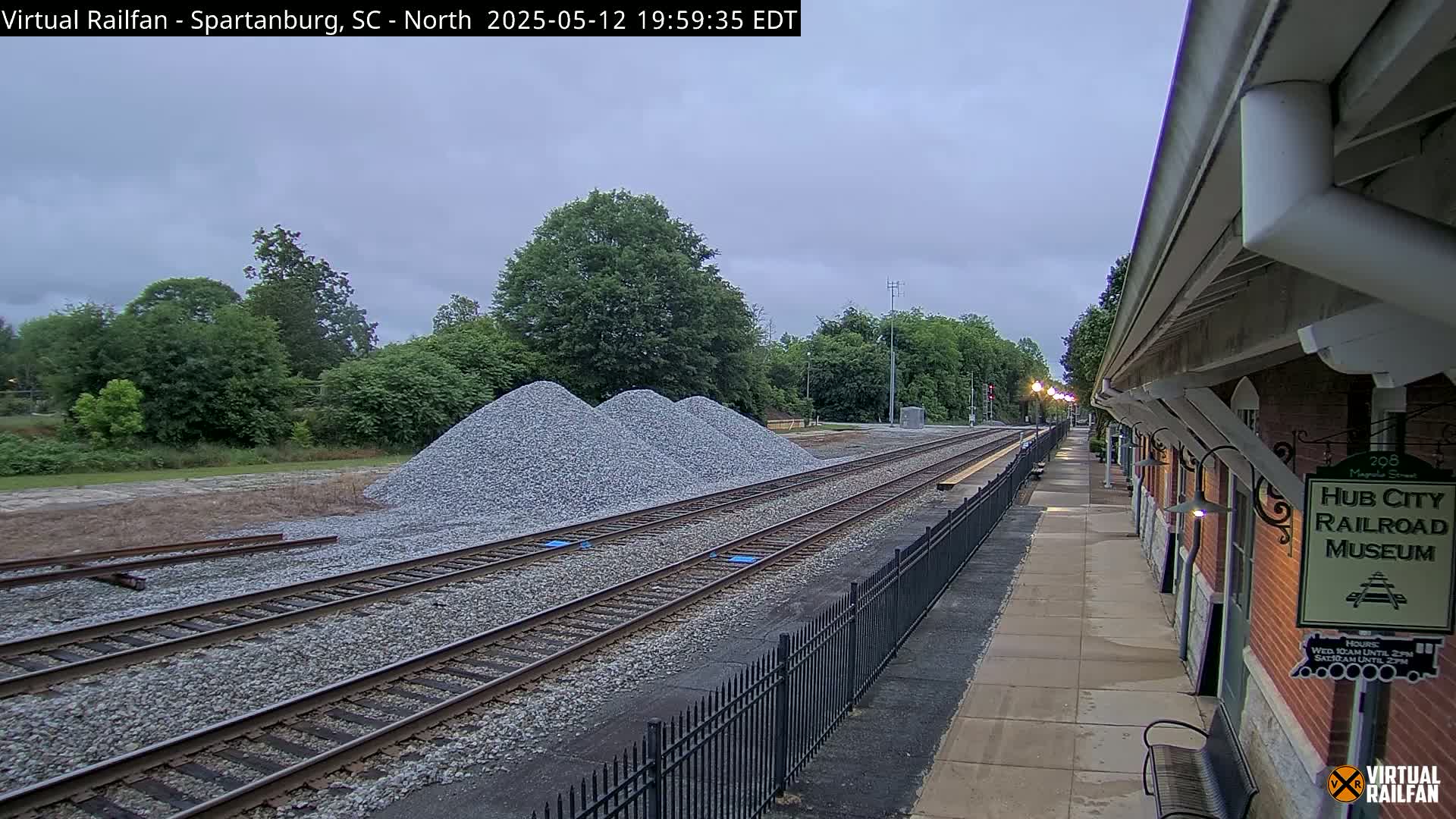 Under an overcast sky, railroad tracks extend from a brick building with several piles of gravel nearby.