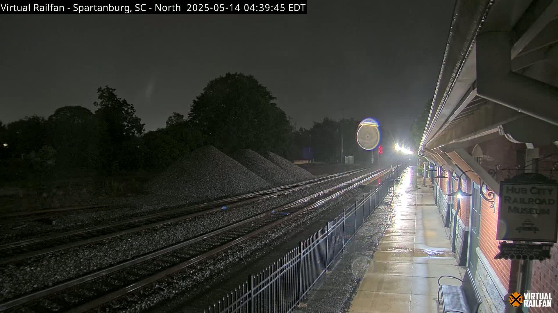 A nighttime view of railroad tracks and a brick building beside them, under rainy conditions.