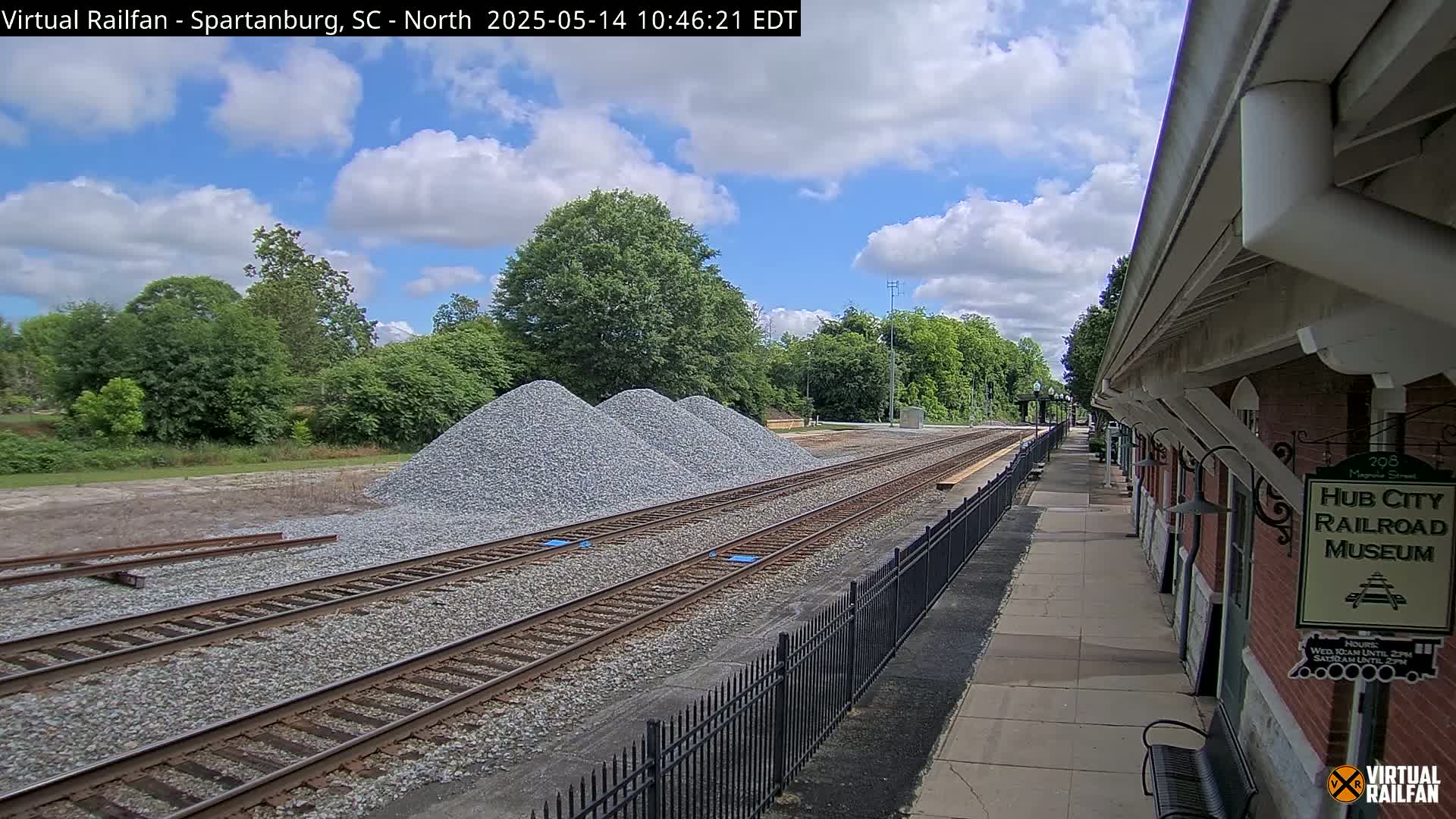 Under a partly cloudy sky, railroad tracks run alongside a brick building, with piles of gravel nearby.
