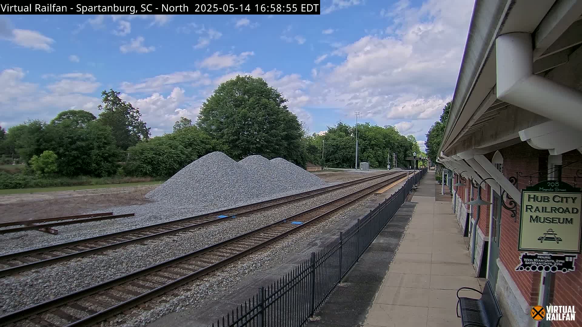 Three piles of gravel sit beside railroad tracks next to a brick building under a partly cloudy sky.