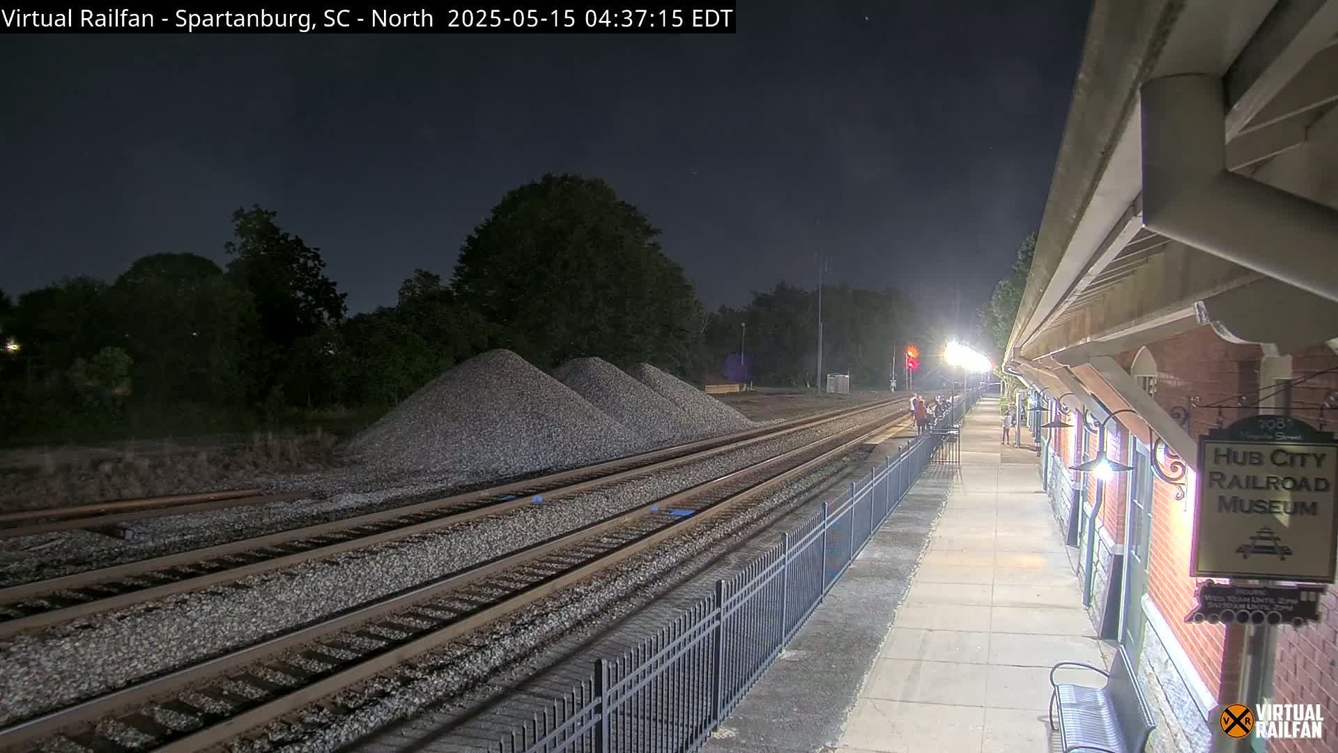 At night, several piles of gravel sit beside a train track that runs alongside a train platform with lights.