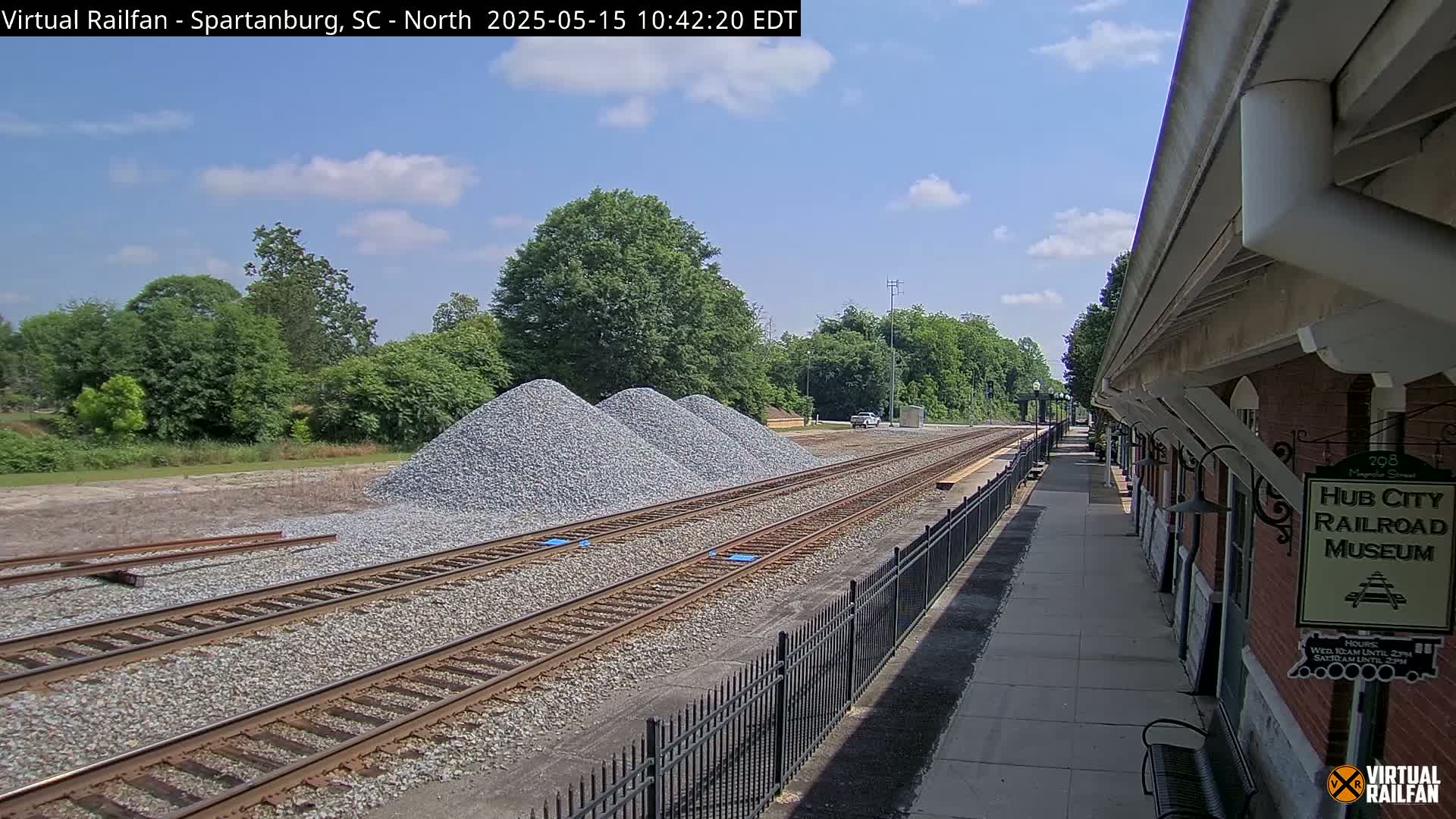 Under a partly cloudy sky, railroad tracks extend from a train platform toward piles of gravel and a line of trees.