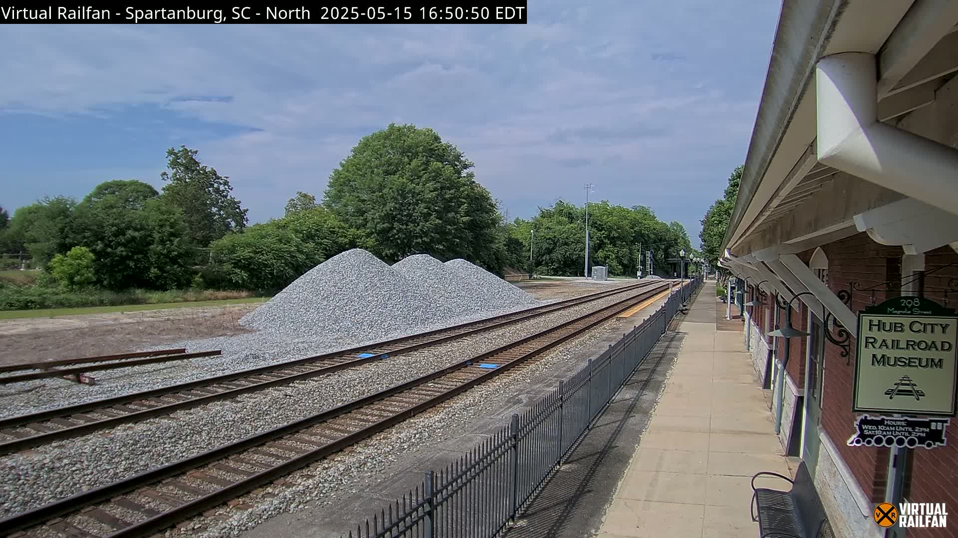 Under a partly cloudy sky, railroad tracks extend from a train platform toward a pile of gravel and trees in the distance.
