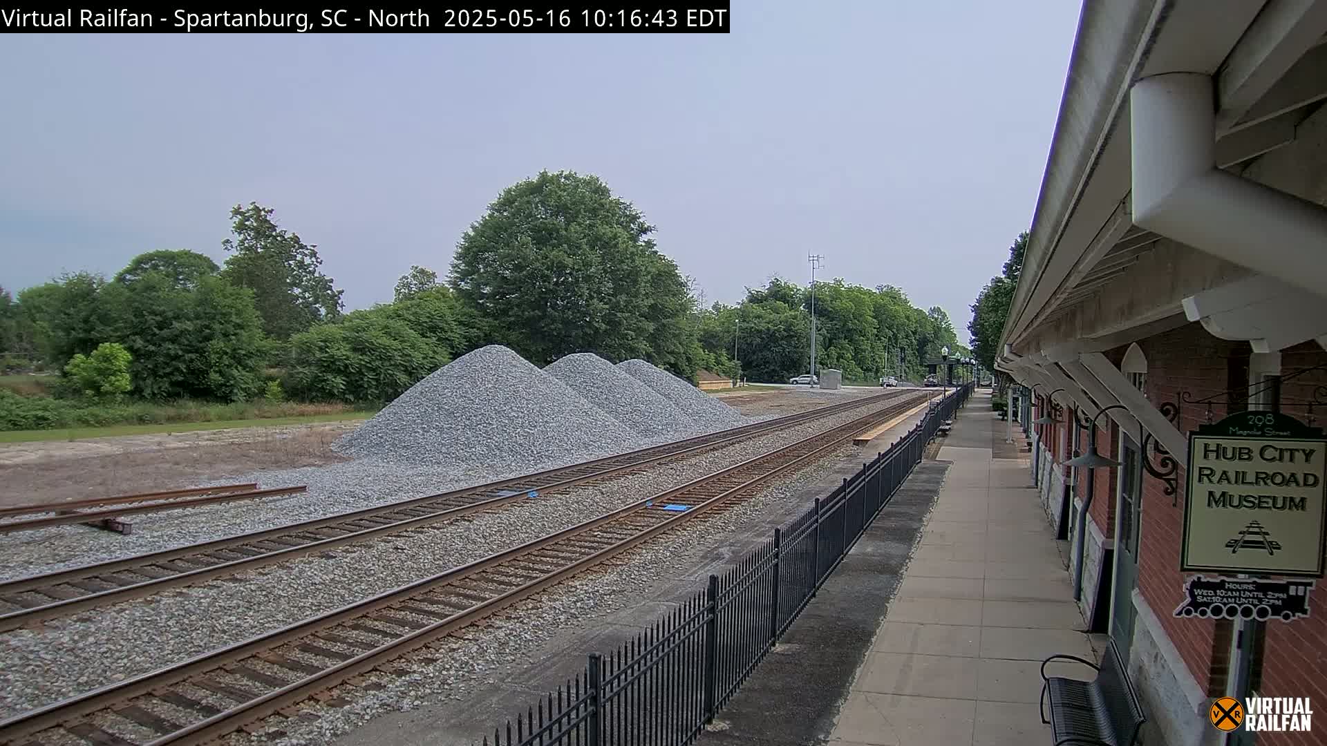 A train platform next to railroad tracks, with piles of gravel nearby, under a partly cloudy sky.