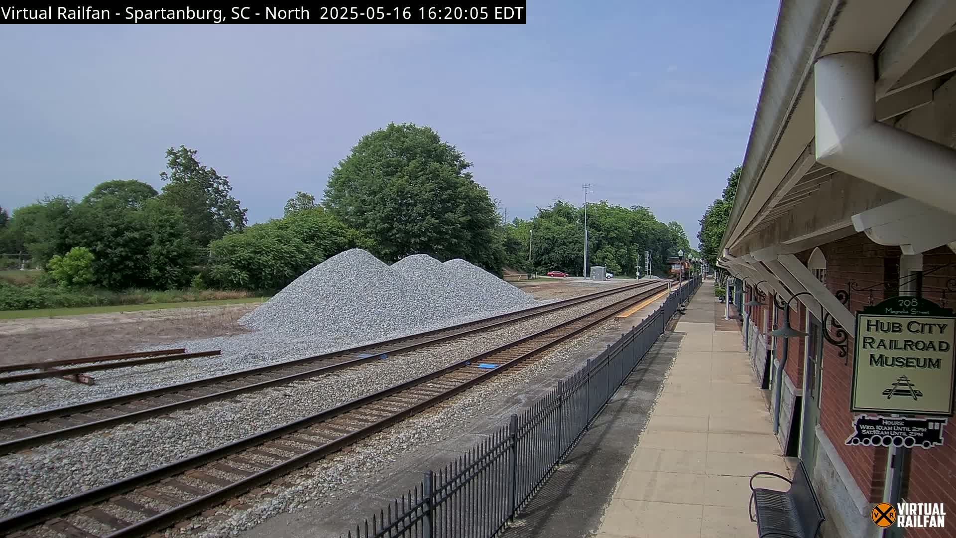 Under a partly cloudy sky, a train platform with a brick building and piles of gravel next to train tracks is visible.