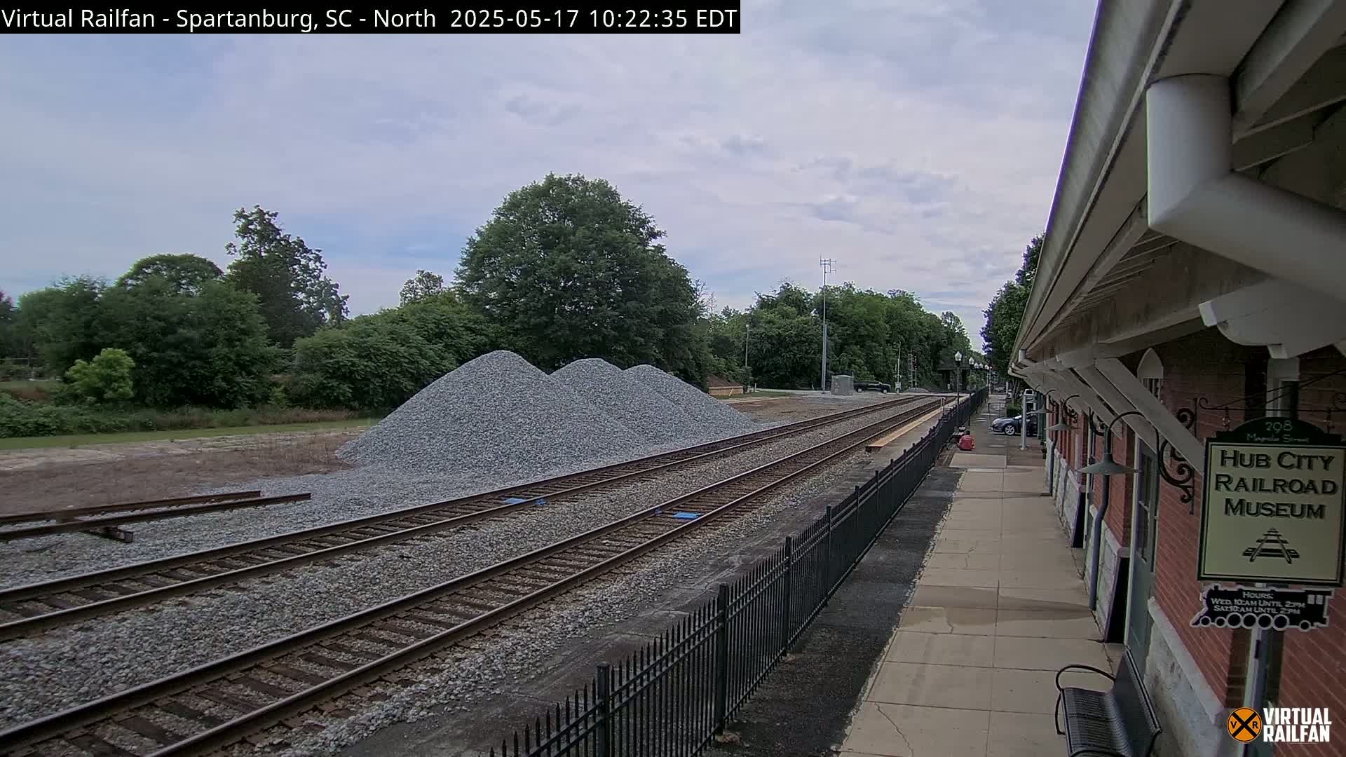 A train track with three piles of gravel next to it runs alongside a brick building under a partly cloudy sky.