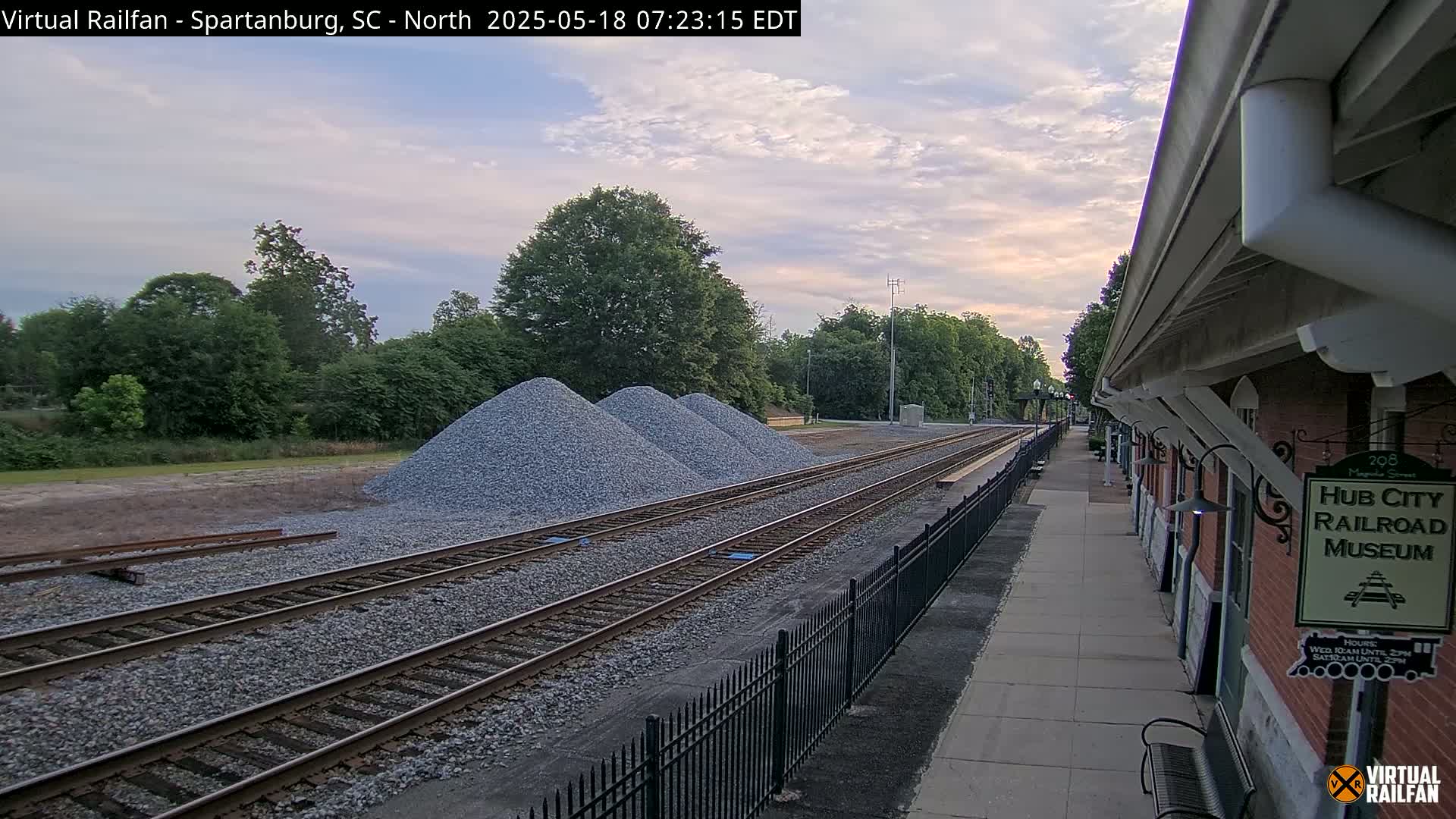 Under a partly cloudy sky, several piles of gravel are situated beside railroad tracks that lead to a brick railroad museum.