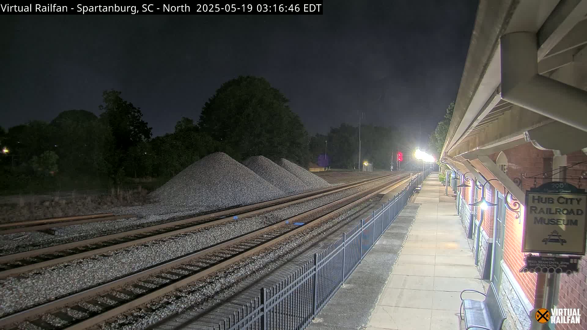 At night, railroad tracks extend from a brick railroad museum toward piles of gravel under a dark sky.