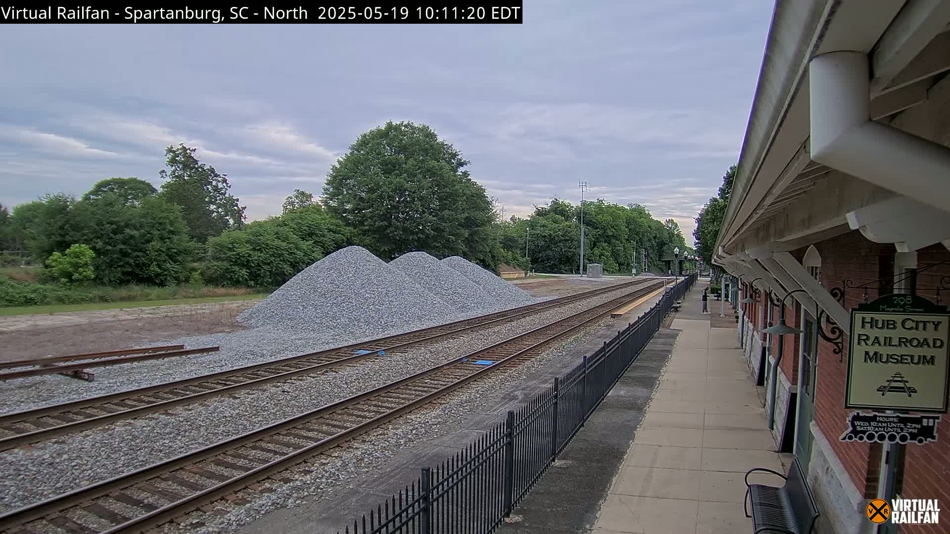 Three piles of gravel are situated beside railroad tracks next to a brick building under a partly cloudy sky.
