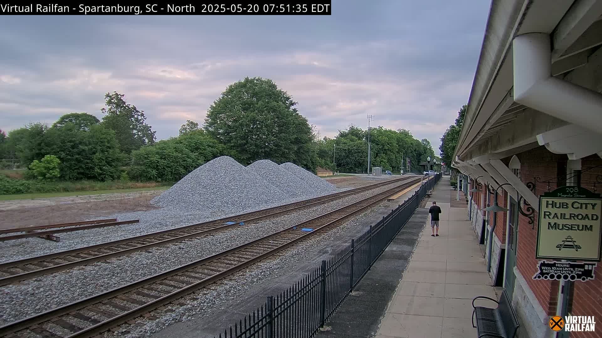 A person walks along a train platform beside railroad tracks with piles of gravel nearby, under an overcast sky.