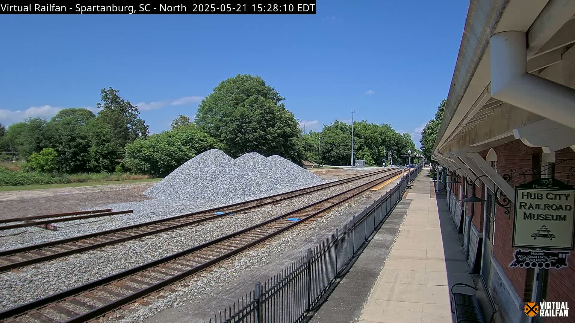 A sunny day shows railroad tracks next to a brick building, with piles of gravel nearby.