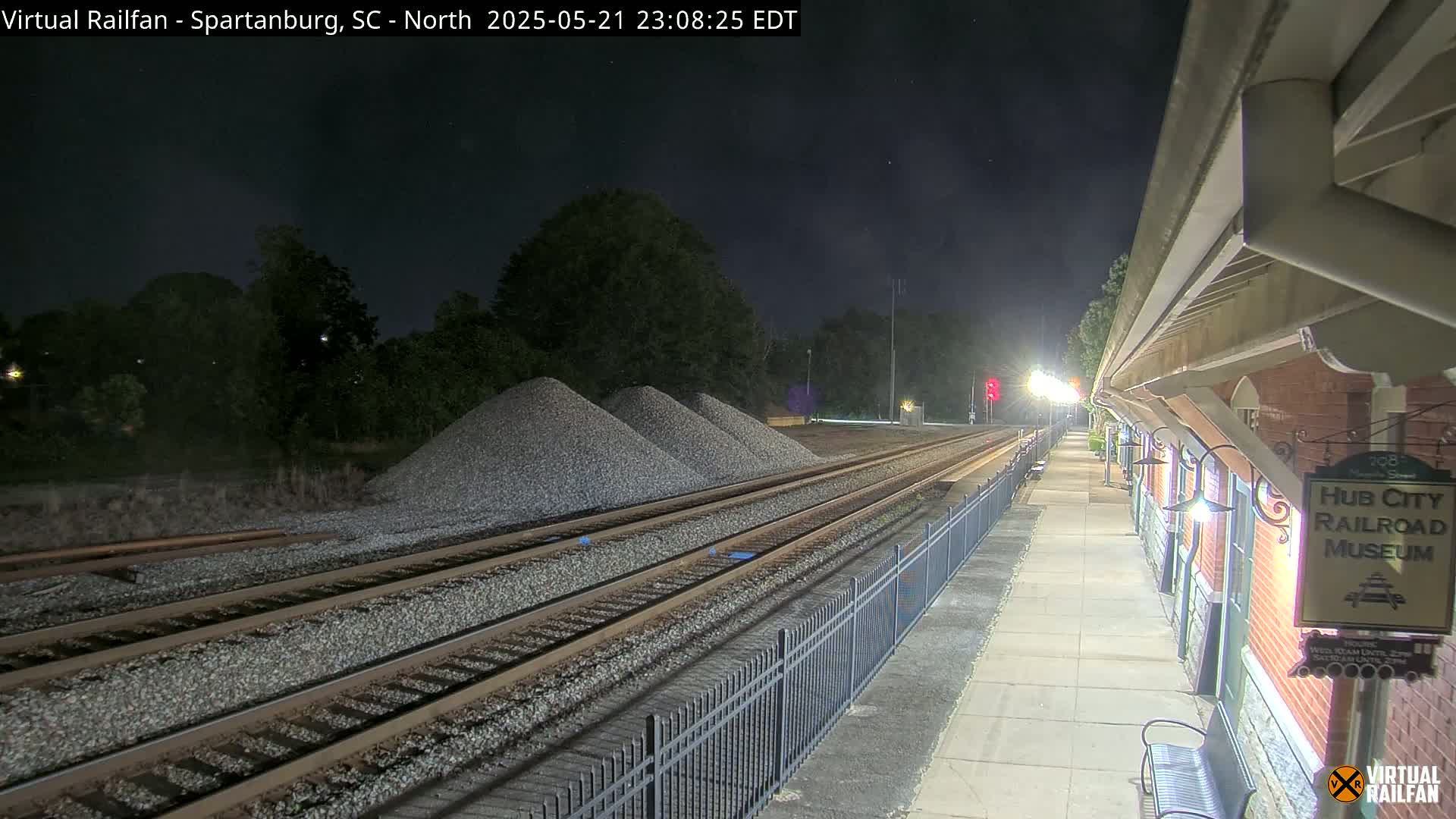 At night, railroad tracks extend from a train platform next to a brick building toward several piles of gravel under a clear, dark sky.